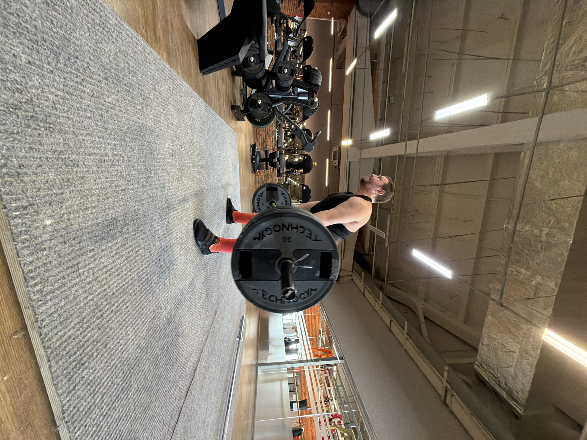 A man is lifting a barbell in a gym. The gym appears to be well-equipped with various weight machines. The location is potentially Chaikovsky, Russia, based on the prompt. The man is wearing athletic attire and appears to be in the middle of a lift. The gym has a gray carpet and the walls have a metal finish and horizontal lighting fixtures.