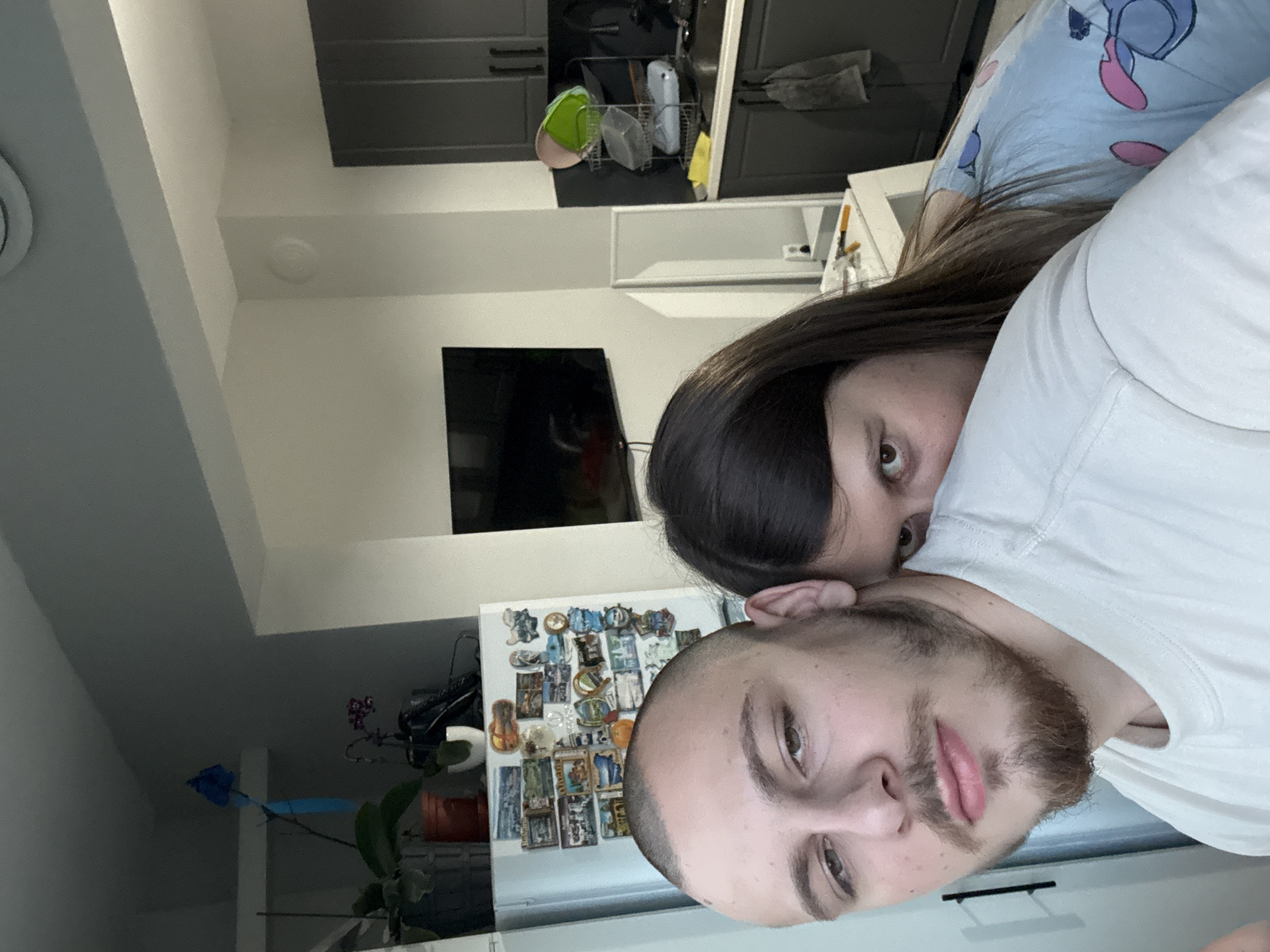A selfie of two people, a male and a female, inside a kitchen. The male has a shaved head and a beard. The female has long, dark hair. Behind them is a refrigerator covered in magnets. Kitchen cabinets are visible in the background. There is a TV on the wall. There is no information available to identify the city of Stara Zagora, Bulgaria from the photo.