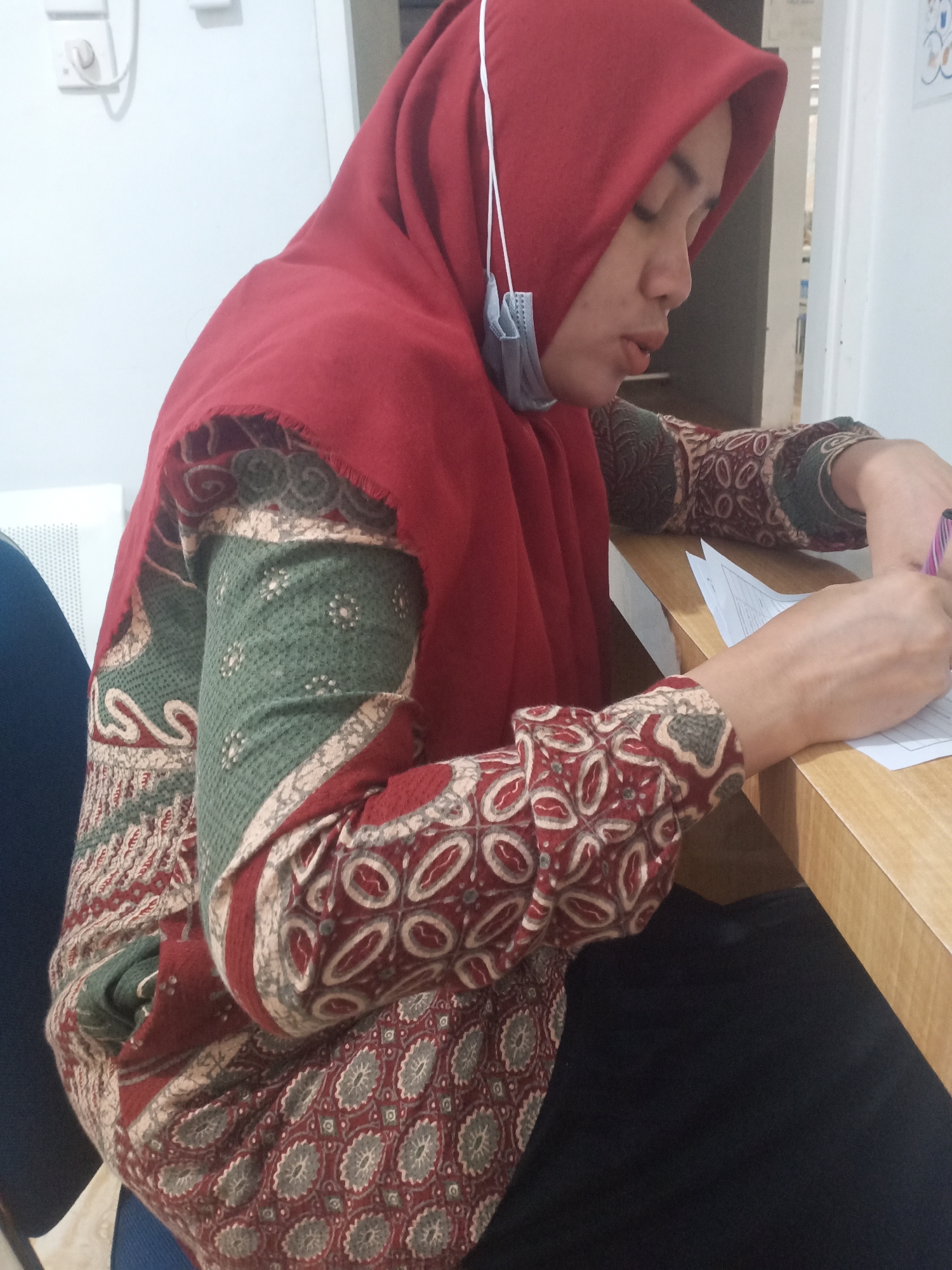 A woman wearing a red hijab and a patterned blouse is seated at a wooden table, writing on a document. She is looking down at the document, and a light blue face mask hangs from the straps of her hijab. The background appears to be an indoor setting, possibly an office or classroom, with a white wall and electrical outlets visible. A white vent is also present on the left side of the image. The woman's hands are visible as she holds a pen and writes. The overall scene is static and focused on the woman's activity.