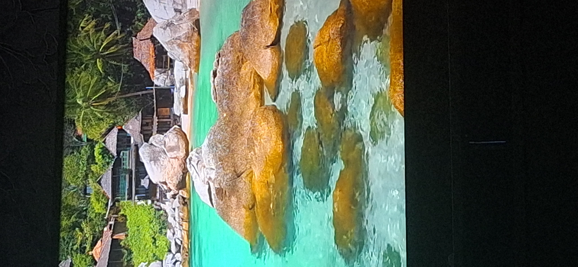This image shows a tropical beach scene with clear turquoise water and large, smooth, tan-colored rocks partially submerged in the water. In the background, there are lush green trees and vegetation, along with several structures with thatched roofs and wooden walls, characteristic of a resort or village in a tropical setting. The overall impression is one of a serene and idyllic coastal environment. The image does not provide any information about the city of Bila Tserkva, Ukraine, nor does it contain any people or specific actions.