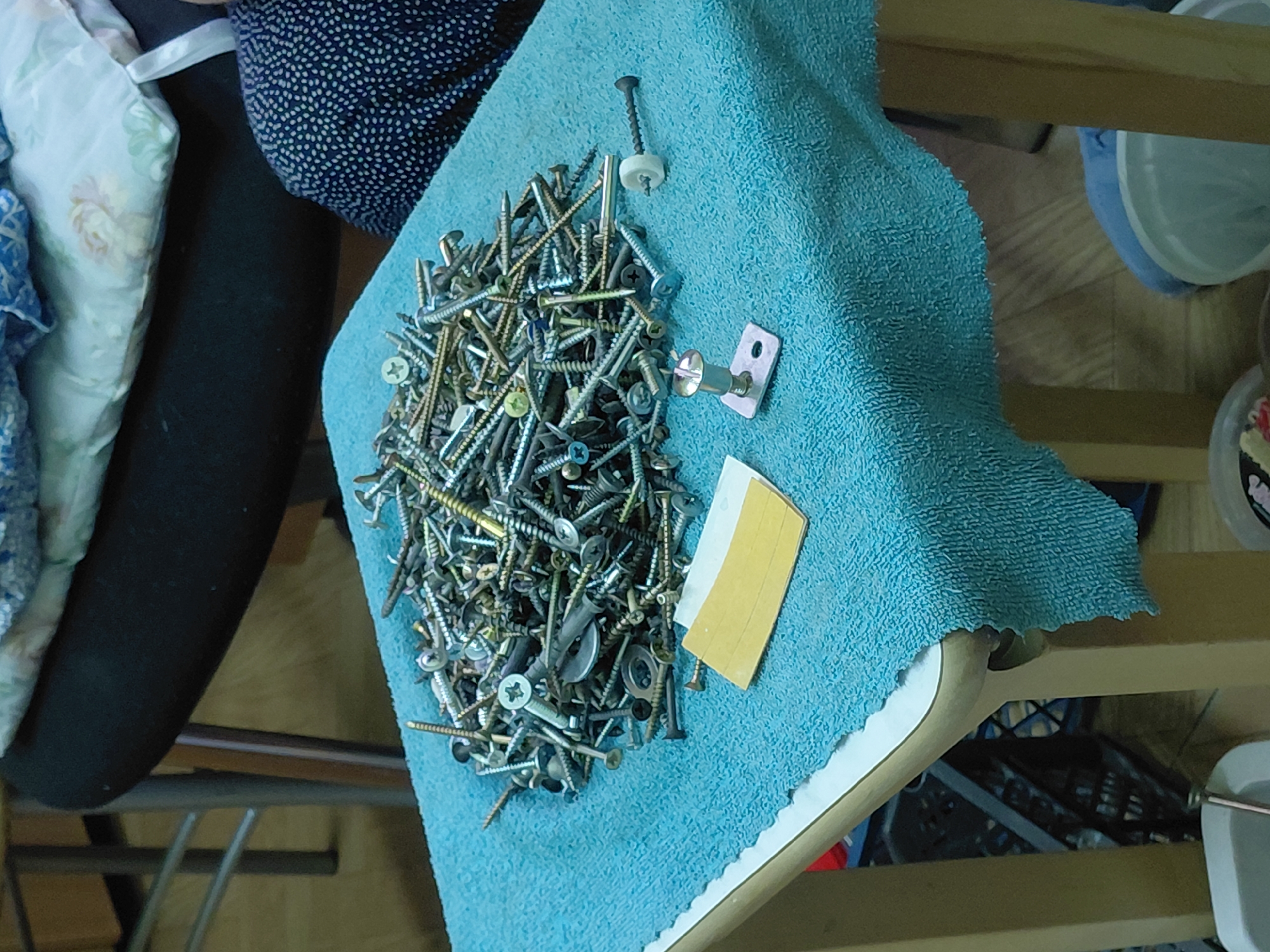 The image shows a close-up of a pile of various screws, a metal bracket, and a yellow piece of paper on a light blue towel. The towel is draped over what appears to be a wooden chair or stool. Parts of a floral-patterned and polka-dot-patterned cushion and chair are visible to the left. A set of shelves with containers on them are visible to the right. The environment is indoors. The image's location is not determined in the image. No people are visible.