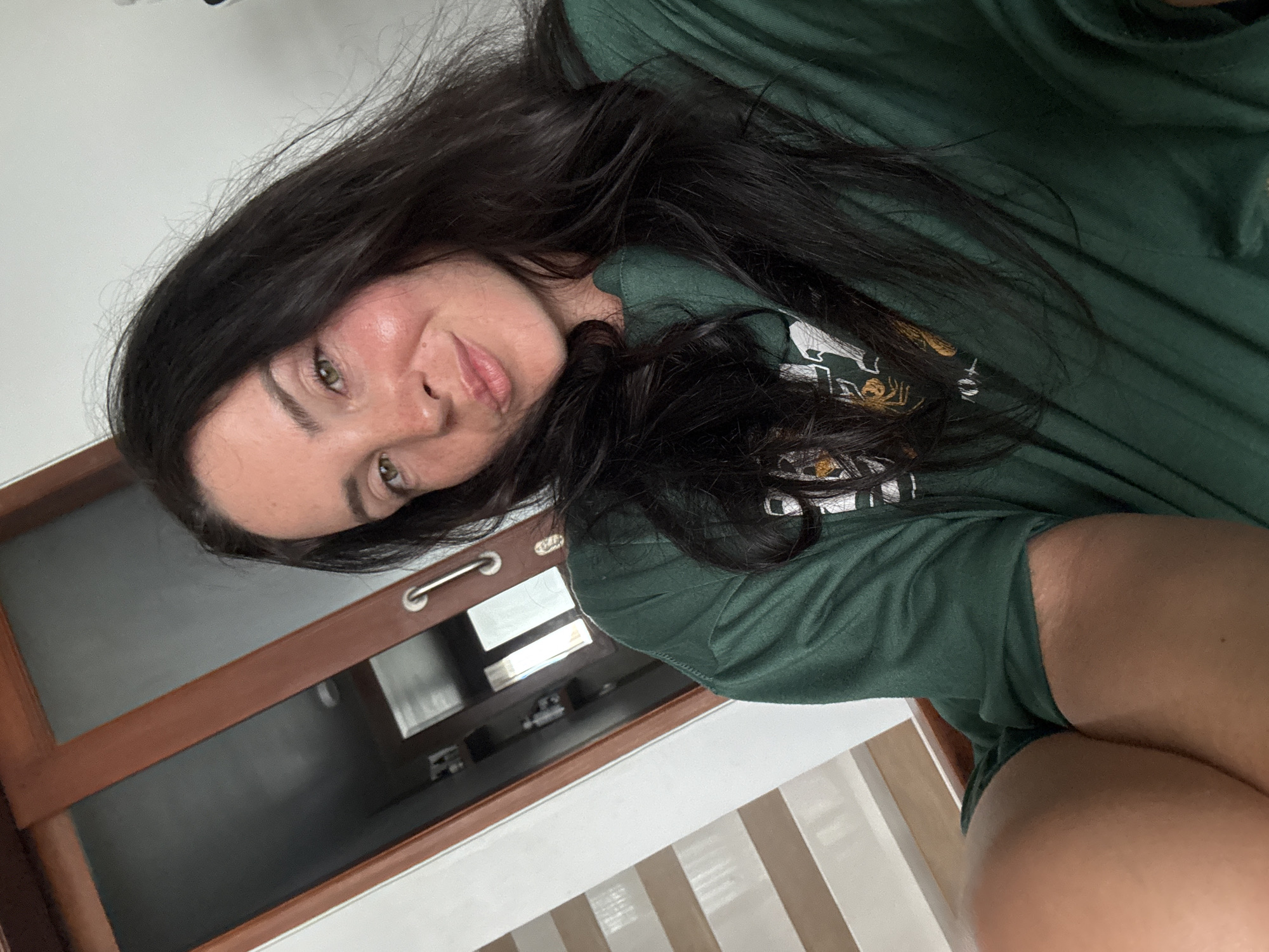 A young woman with long, dark hair is posing for a selfie indoors. She is wearing a green t-shirt. The background includes a wooden door, a staircase, and a wall. The scene appears to be a residential interior.