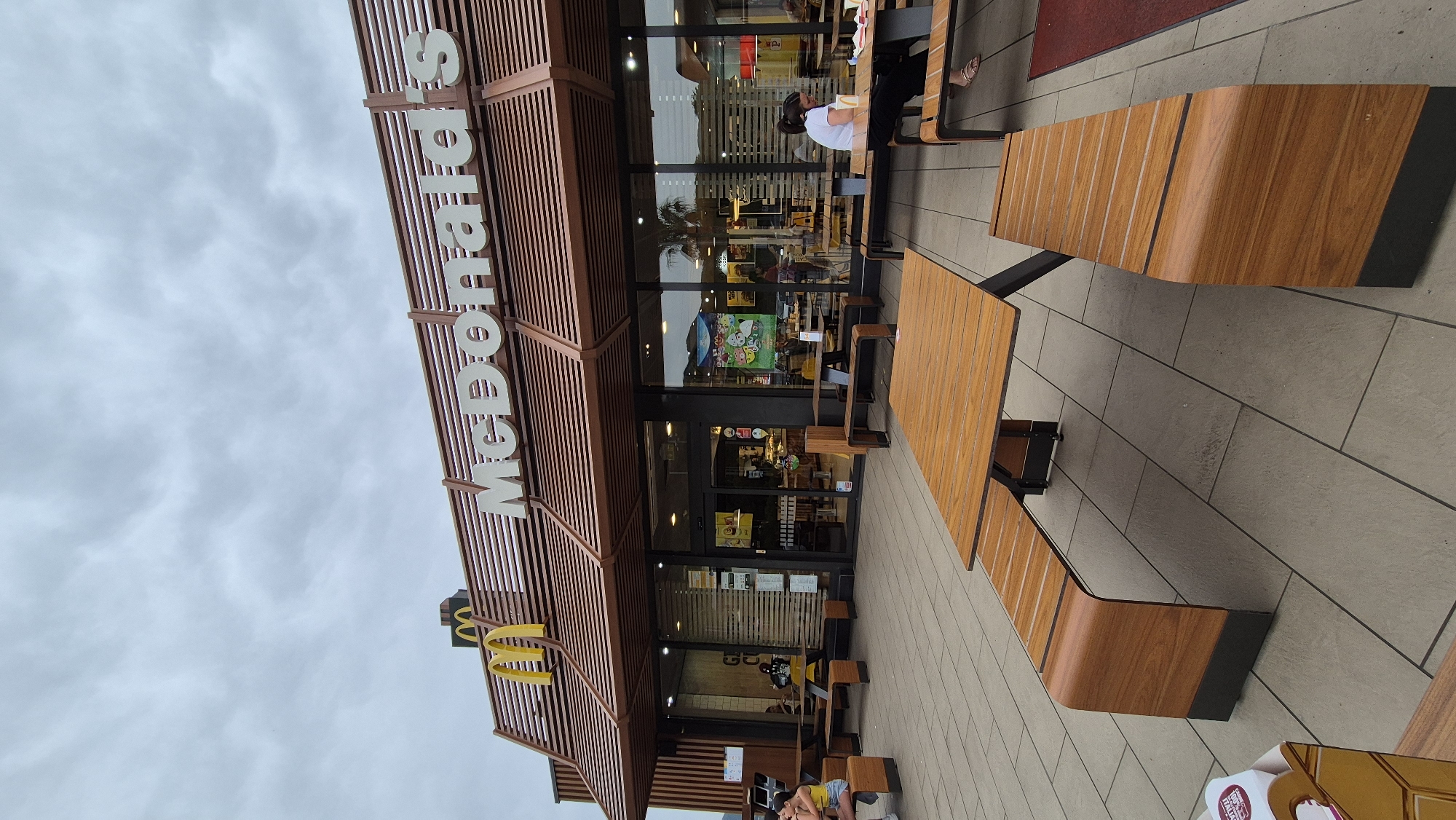 An exterior, medium shot captures a McDonald's restaurant with an outdoor seating area on a cloudy day. The restaurant features a modern facade with a brown slatted roof and large glass windows, displaying the "McDonald's" logo in white lettering and the iconic golden arches. Inside, the restaurant is dimly lit, with some patrons visible. Outside, several wooden picnic tables and benches are arranged on a tiled patio. Two young women are seated at one of the tables on the left, and a woman is seated at a table on the right. The sky is overcast with gray clouds.