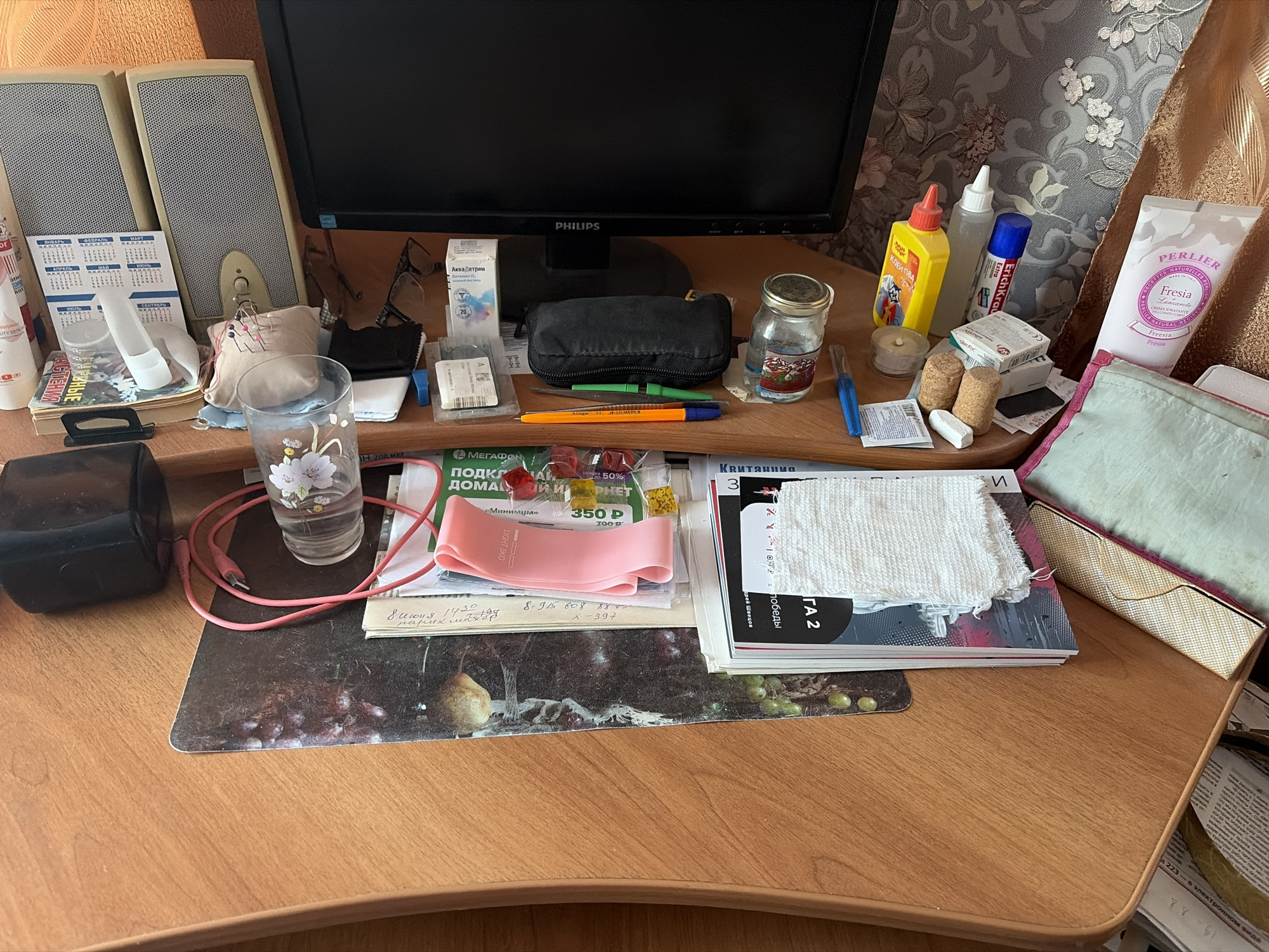 The image depicts a desk surface with various items, including a computer monitor, speakers, pens, a jar, a tube of cream, and other small objects. The items are arranged in a somewhat cluttered manner, suggesting a workspace or study area. The background features a patterned wallpaper and a gold-colored fabric. The overall impression is that of a personal desk with everyday objects. Based on the provided context, this desk is located in Skopin, Russia.