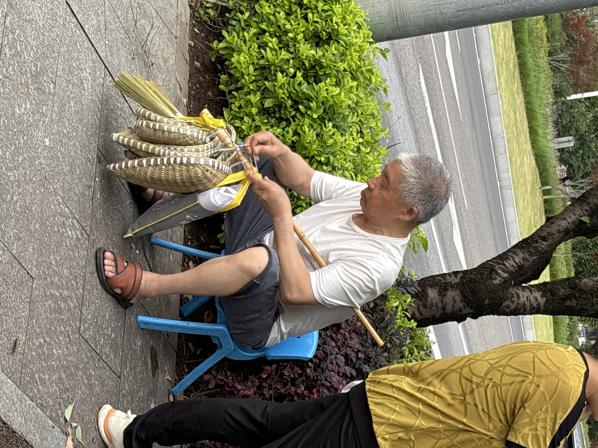 A gray-haired man in a white t-shirt and gray shorts sits on a blue plastic chair on a paved surface next to a planter with green foliage. He appears to be tying a bundle of woven baskets together with yellow ribbon and twine. A second individual wearing black pants and a yellow shirt is partially visible in the lower right corner of the image. A street with white lines and grass medians is visible in the background. The setting is likely urban, potentially Jiangjin, China, based on the request.
