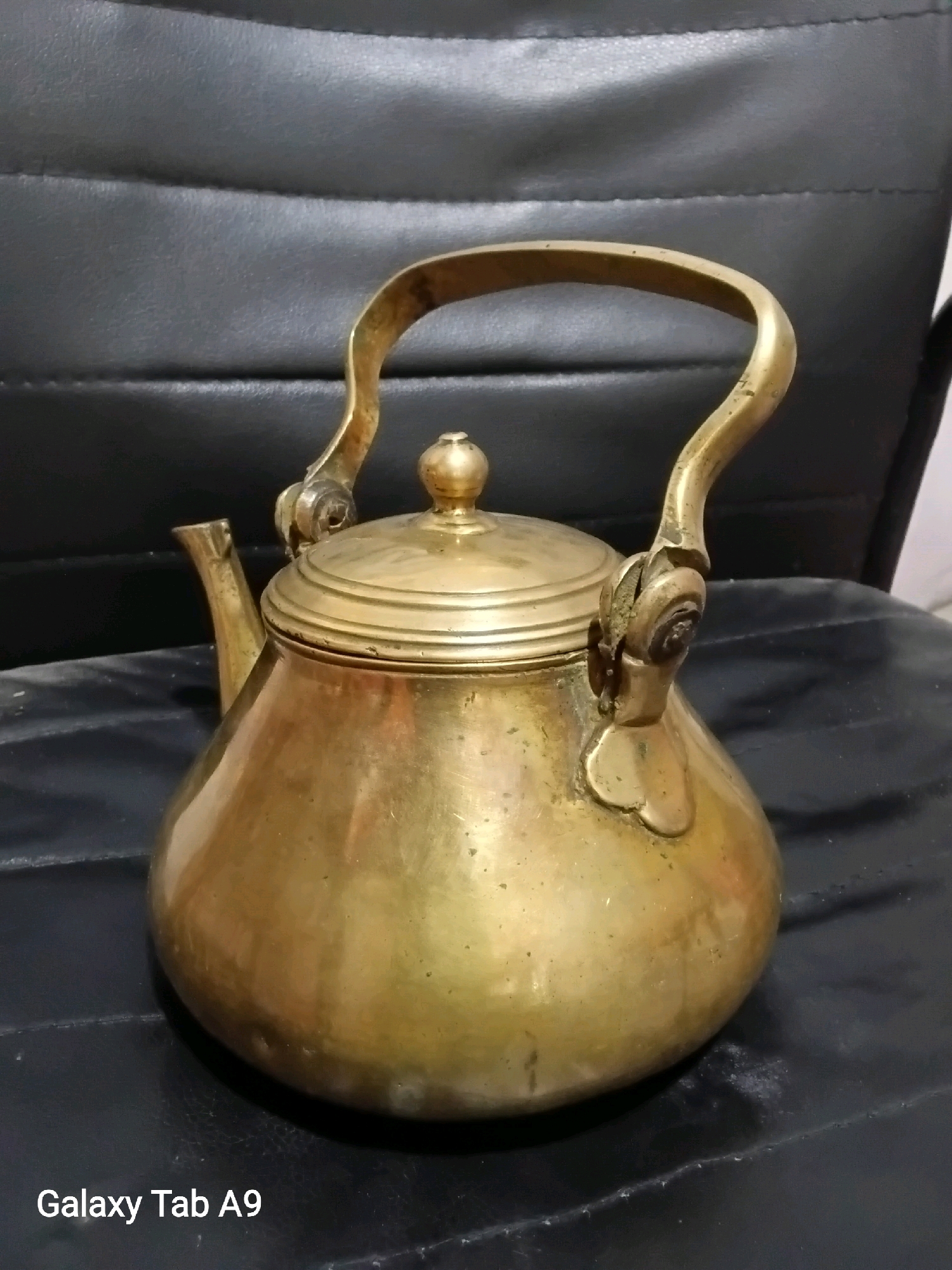 A brass teapot rests on a black, textured surface, likely a chair or couch. The teapot is round with a spout and a curved handle, and has a lid topped with a small knob. The brass exhibits some signs of age and use, with minor discoloration and a slightly dulled sheen. The background is dark and indistinct. The image has a watermark in the lower left corner that reads "Galaxy Tab A9". There is no information about the location of Jati, Indonesia.