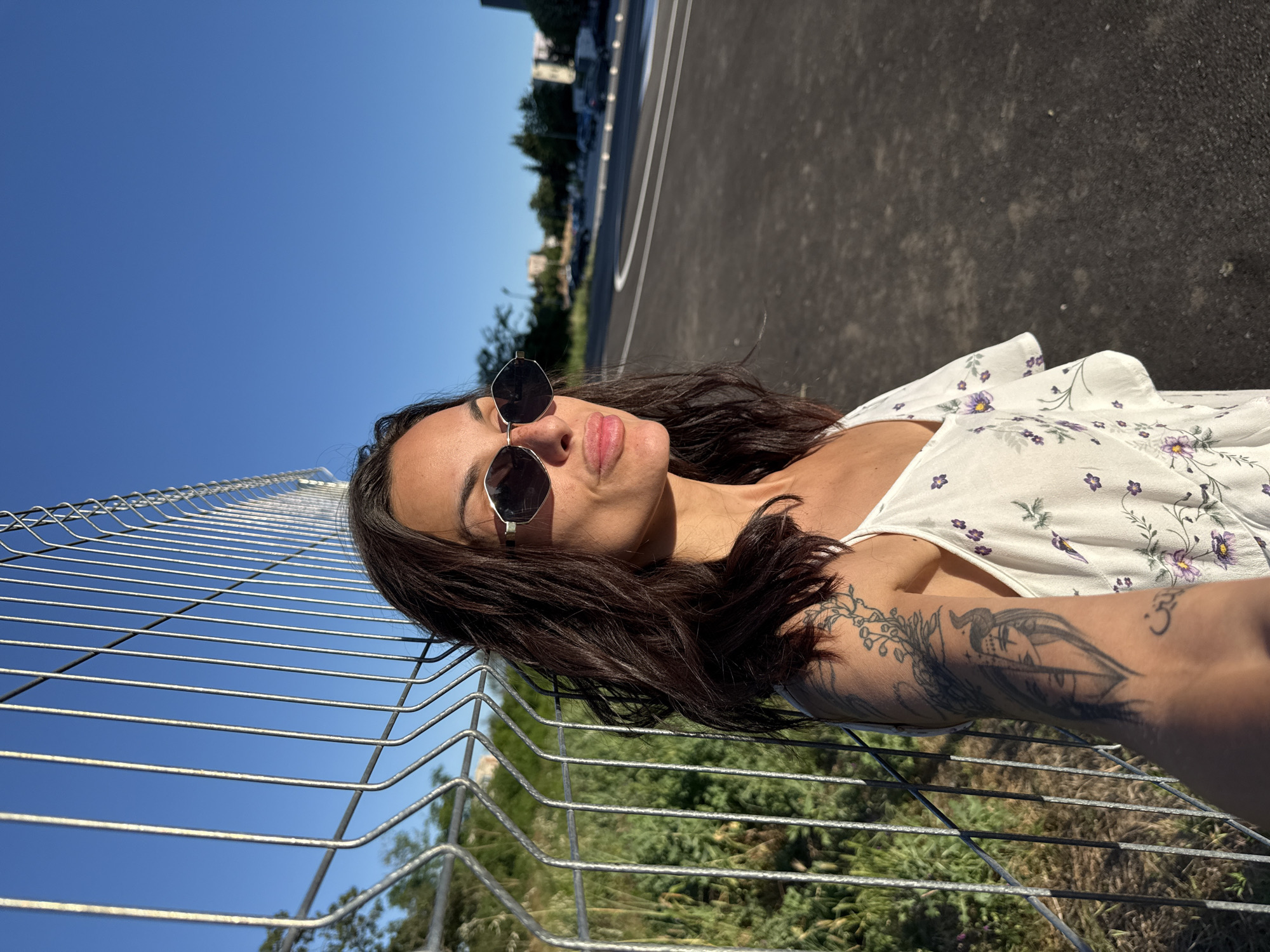 In the city of Pleven, Bulgaria, a woman with dark hair and sunglasses stands near a chain-link fence. She is wearing a white floral dress. Her left arm is visible and features several tattoos, including what appears to be a face with horns and swirling designs. The background consists of a clear blue sky, green vegetation, and a paved area with markings. The lighting suggests it is late afternoon or early evening.