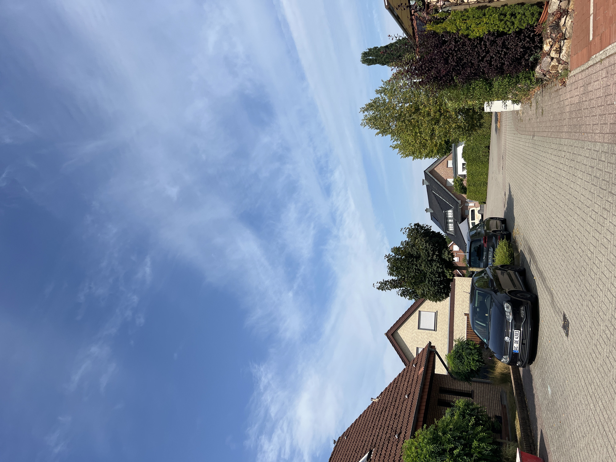 A street view shows two parked cars, a black Volkswagen Polo and a dark gray SUV, on a paved driveway in front of residential houses. The Volkswagen Polo is in the foreground, parked on the left side of the driveway, and its license plate is visible. The houses are of brick construction with red tile roofs. Trees and landscaping are present on either side of the driveway. The sky is blue with wispy white clouds. The scene depicts a quiet suburban street on a clear day.