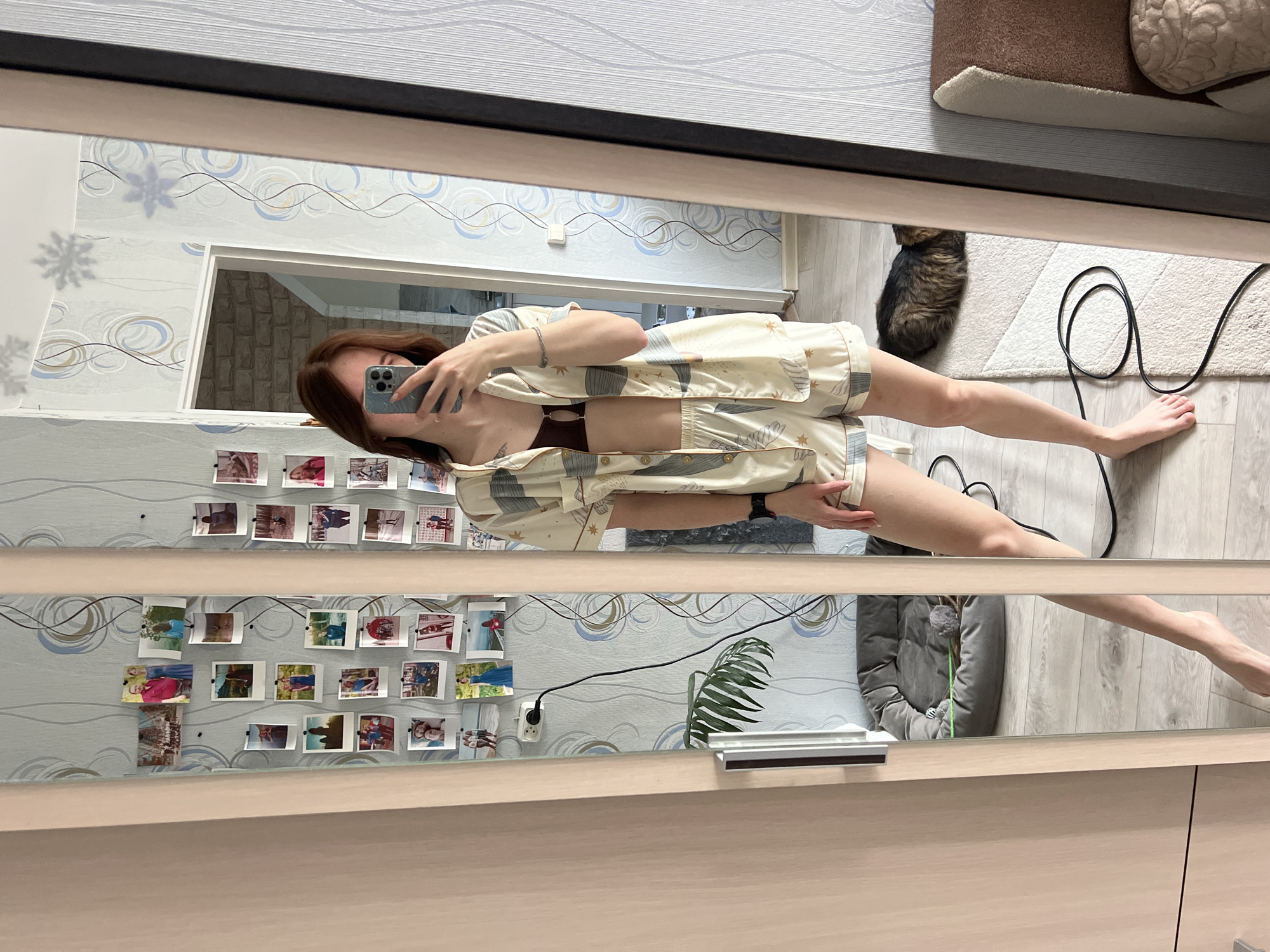 This image is a full-length mirror selfie, presented with a 90-degree clockwise rotation, taken indoors in Chaikovsky, Russia. The reflection shows a person, likely a young woman with long, dark brown hair, holding a smartphone to capture the picture. She is casually dressed in a light-colored pajama set featuring a pattern of abstract shapes, possibly including stars or celestial motifs, worn over a dark brown bra. Her face is partially obscured by the phone as she holds it up.
The setting appears to be a bedroom or dressing area with light wooden flooring and wallpaper that features a subtle blue and grey wavy or swirling pattern. Several small, unframed photographs are taped or pinned to the wall in multiple rows, displaying various personal snapshots. Snowflake-shaped cutouts are also visible on the wall's surface in the reflection.
In the background, a tabby cat is visible, curled up and resting on a white rug on the floor. A black power cord snakes across the floor, connecting to a white electrical outlet mounted on the patterned wall. A green leafy plant, possibly a palm frond, is reflected near the bottom of the mirror, adjacent to a grey, cushioned pet bed or similar item. The large mirror, likely part of a wardrobe, reflects the entire casual indoor environment. The time of day appears to be daytime due to the general lighting. No visible text is clearly legible in the image.
