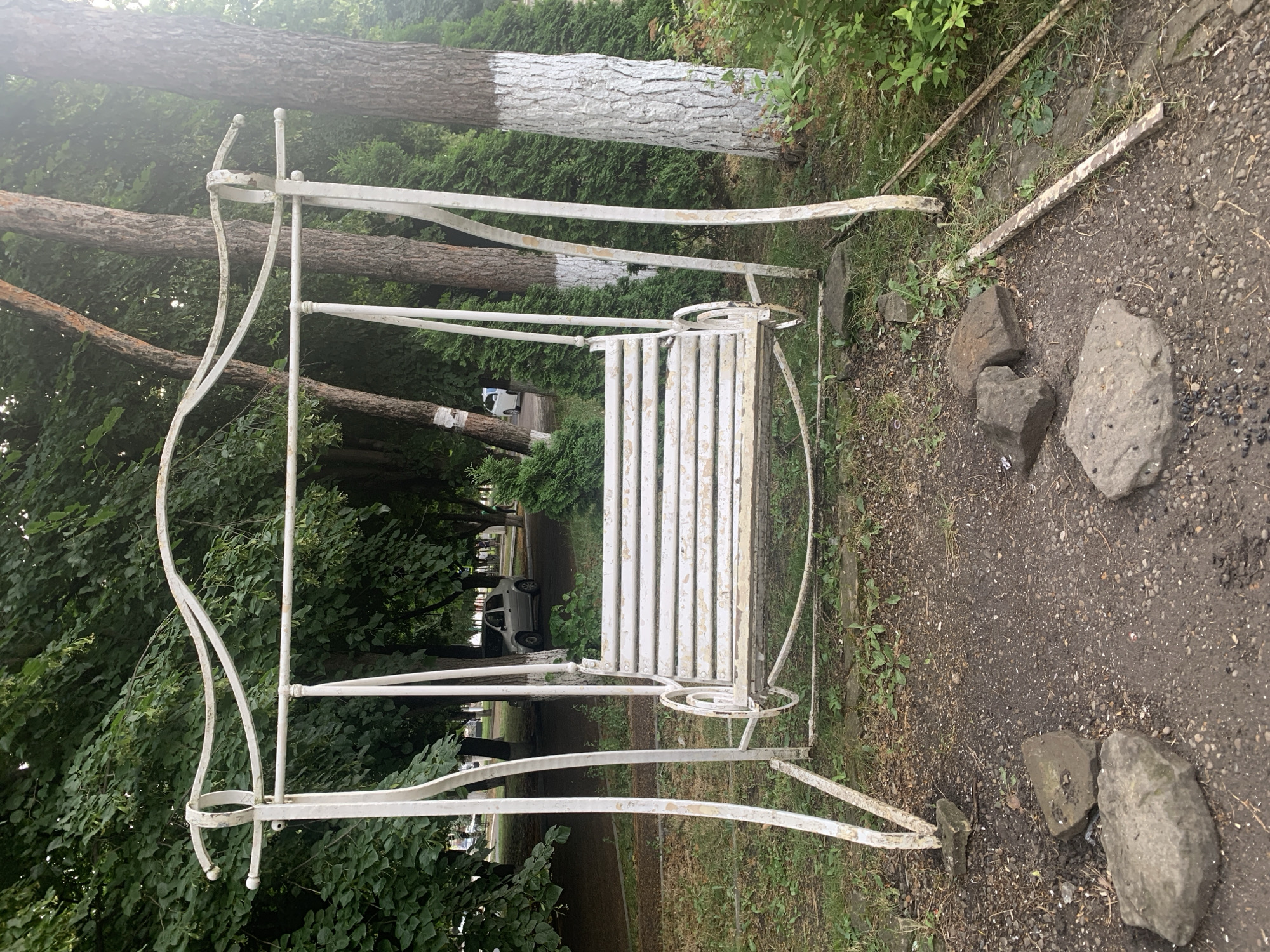An outdoor scene in Dondușeni, Moldova, features a weathered white metal garden swing set with a slatted seat. The swing is situated on a patch of ground covered with a mixture of dirt, gravel, grass, and scattered rocks. Behind the swing, a line of trees with rough bark can be seen, some of which have their lower trunks painted white. Further in the background, more trees and foliage create a dense green backdrop. A light-colored car is visible parked on a paved area among the trees. The overall impression is one of a slightly neglected park or public space.
