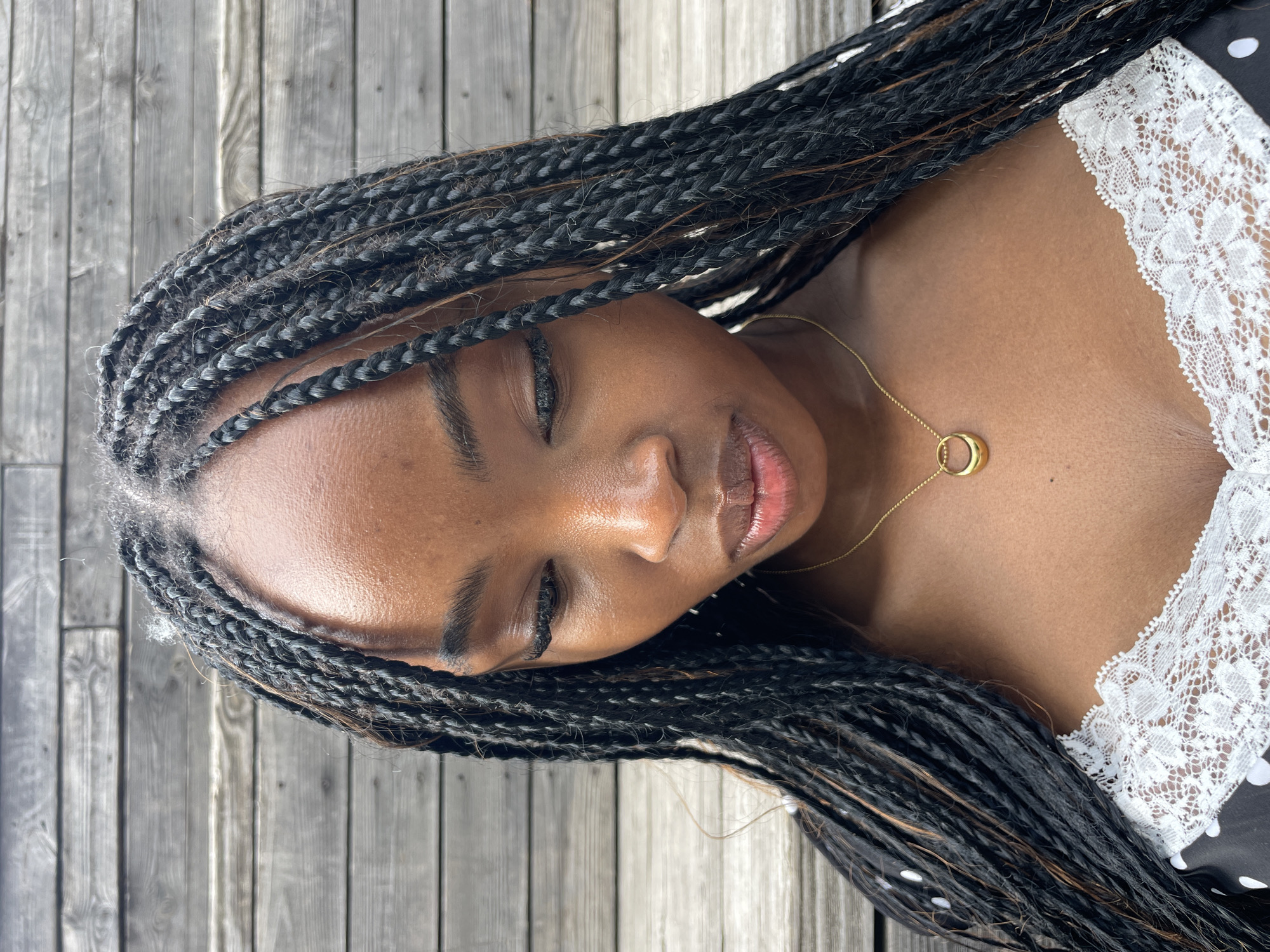 A close-up, head and shoulders portrait captures a young Black woman with braided hair looking down. She wears a gold pendant necklace and a black and white polka dot dress with white lace trim. The background consists of weathered wooden planks, suggesting an outdoor setting. The image does not provide any information that would indicate a location in Kazan, Russia.