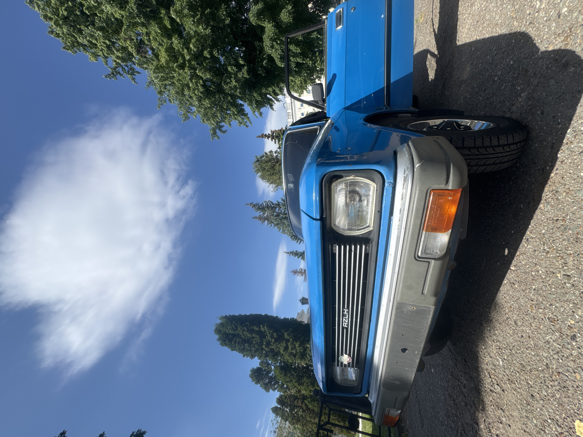 A front-facing view of a blue AZLK car parked on an asphalt surface is shown under a blue sky with white clouds and scattered trees. The car features a gray bumper, rectangular headlight, orange turn signal, and a vertical chrome grill with an AZLK emblem. The driver's side door is open. The location is Kazan, Russia.
