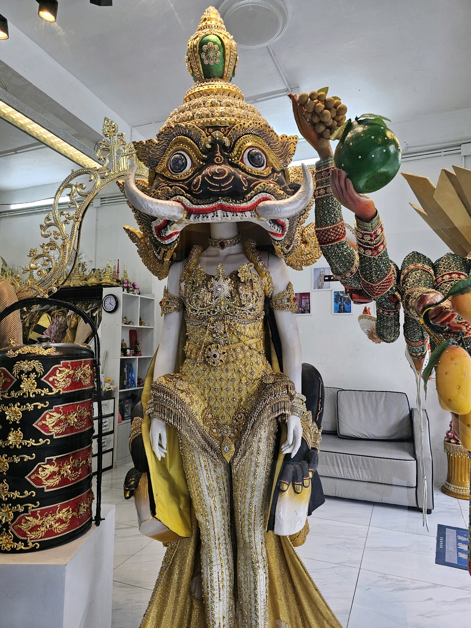 The image depicts a close-up of an elaborate, golden costume displayed on a mannequin in a well-lit room. The costume features a large, ornate headpiece with intricate details, large tusks, and prominent eyes. The body of the costume is heavily embellished with gold fabric, beads, and dangling ornaments. The mannequin is positioned in what appears to be a workshop or studio, indicated by the presence of shelves with various items, a clock on the wall, and a couch in the background. To the right, another figure, partially visible and also adorned in a detailed costume, holds fruit in its hands. The overall impression is one of cultural artistry and craftsmanship, likely related to traditional Thai performances or festivals, possibly located in Bangkok, Thailand, given the context of such elaborate creations.
