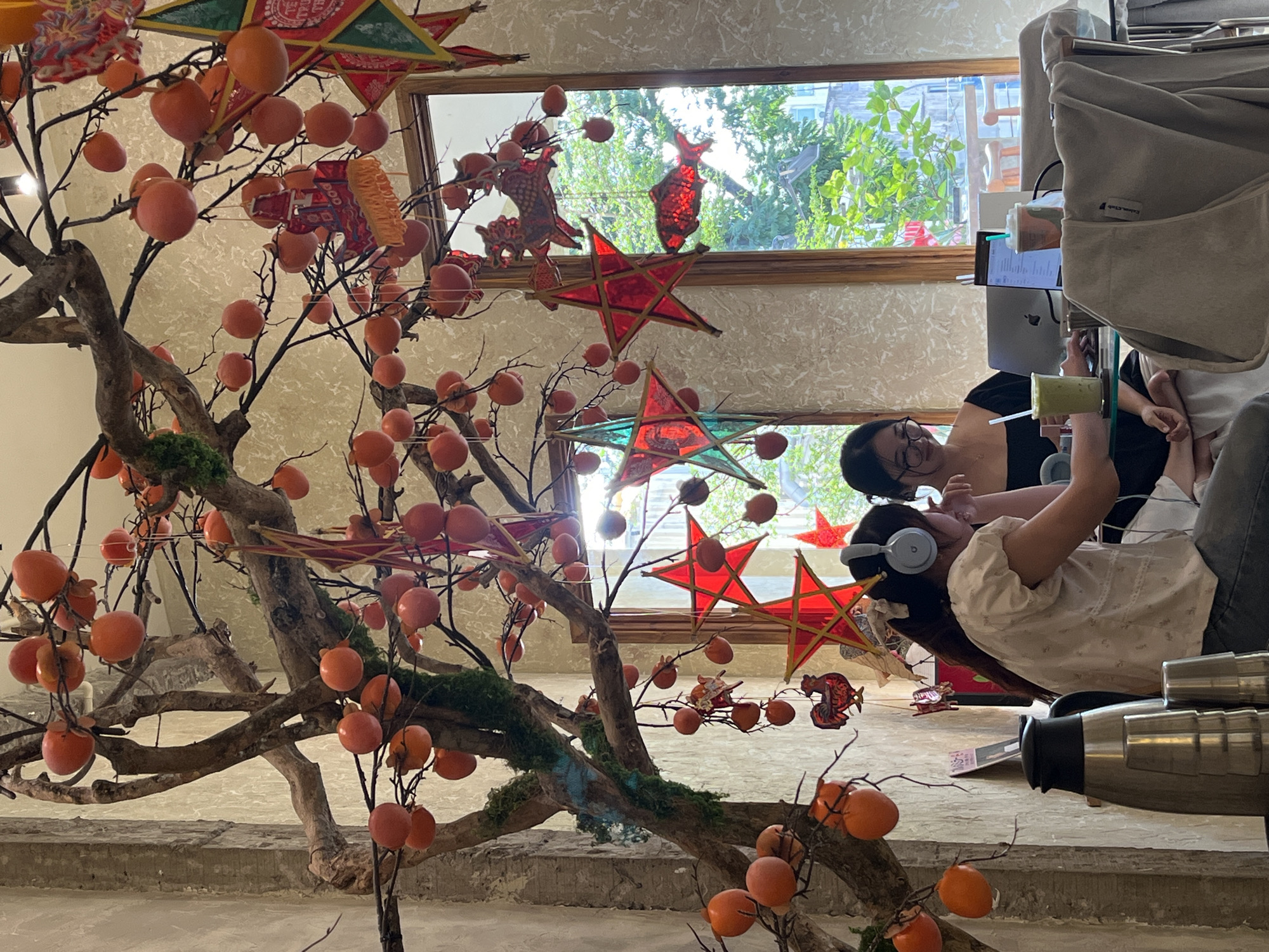 Two young Vietnamese women are seated at a table in a cafe, likely in Ho Chi Minh City, Vietnam, suggested by the style of decoration and the overall ambiance. The cafe is decorated with a large, artificial tree adorned with numerous persimmons and various festive lanterns, including red stars and what appears to be a traditional Vietnamese fish lantern.
The woman on the left, wearing headphones, has long dark hair and is dressed in a white shirt. She is looking towards her right, possibly at the other woman or something beyond the frame. The woman on the right, wearing glasses, has dark hair pulled back and is wearing a black off-the-shoulder top. She is looking down at a laptop and appears to be working or studying. A green beverage with a straw is in front of her, along with a laptop and another drink.
In the foreground, a metallic thermal carafe and several metal cups are stacked, suggesting a self-service beverage station or part of the cafe's display. The background shows a large window with lush green trees and foliage visible outside, indicating a bright, possibly daytime setting. The wall behind the tree has a textured, neutral finish. The scene depicts a casual, relaxed atmosphere, likely a common sight in a modern cafe in Ho Chi Minh City catering to students or individuals seeking a workspace.