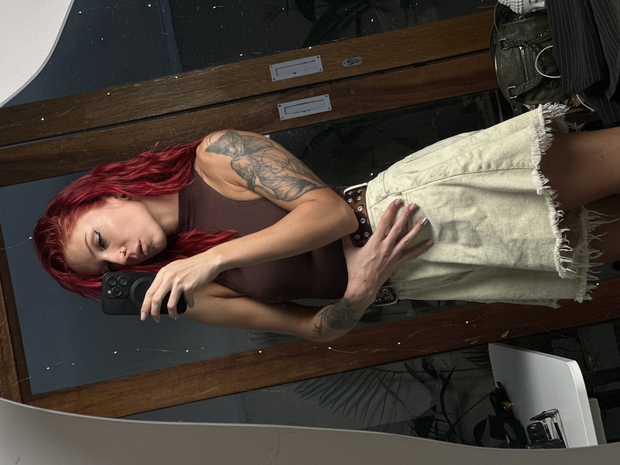 A mirror selfie captures a young woman with vibrant red, wavy hair. She is wearing a fitted brown ribbed tank top and a light beige or off-white denim mini-skirt with a frayed hem, accentuated by a dark brown belt with metal eyelets. Her left arm, holding a black smartphone with a MagSafe ring, is visible. Large, detailed tattoos adorn her upper arms, and another is on her right forearm. She has a nose piercing, light eye makeup, and manicured nails, with one appearing silver and others light pink.
The woman is posing at an angle, looking towards her reflection with a neutral to slightly contemplative expression. The mirror reflects an indoor setting featuring dark surfaces, possibly wooden door frames or panels, and faint string lights. Other elements in the reflection include what appears to be foliage from a plant, a dark green or brown leather bag with metallic accents, a striped piece of clothing, and a white electronic device resembling a router or monitor stand. The lighting suggests an indoor environment, potentially in the evening or under low light. No text is visible in the image. The scene takes place in North Kuta, Indonesia.