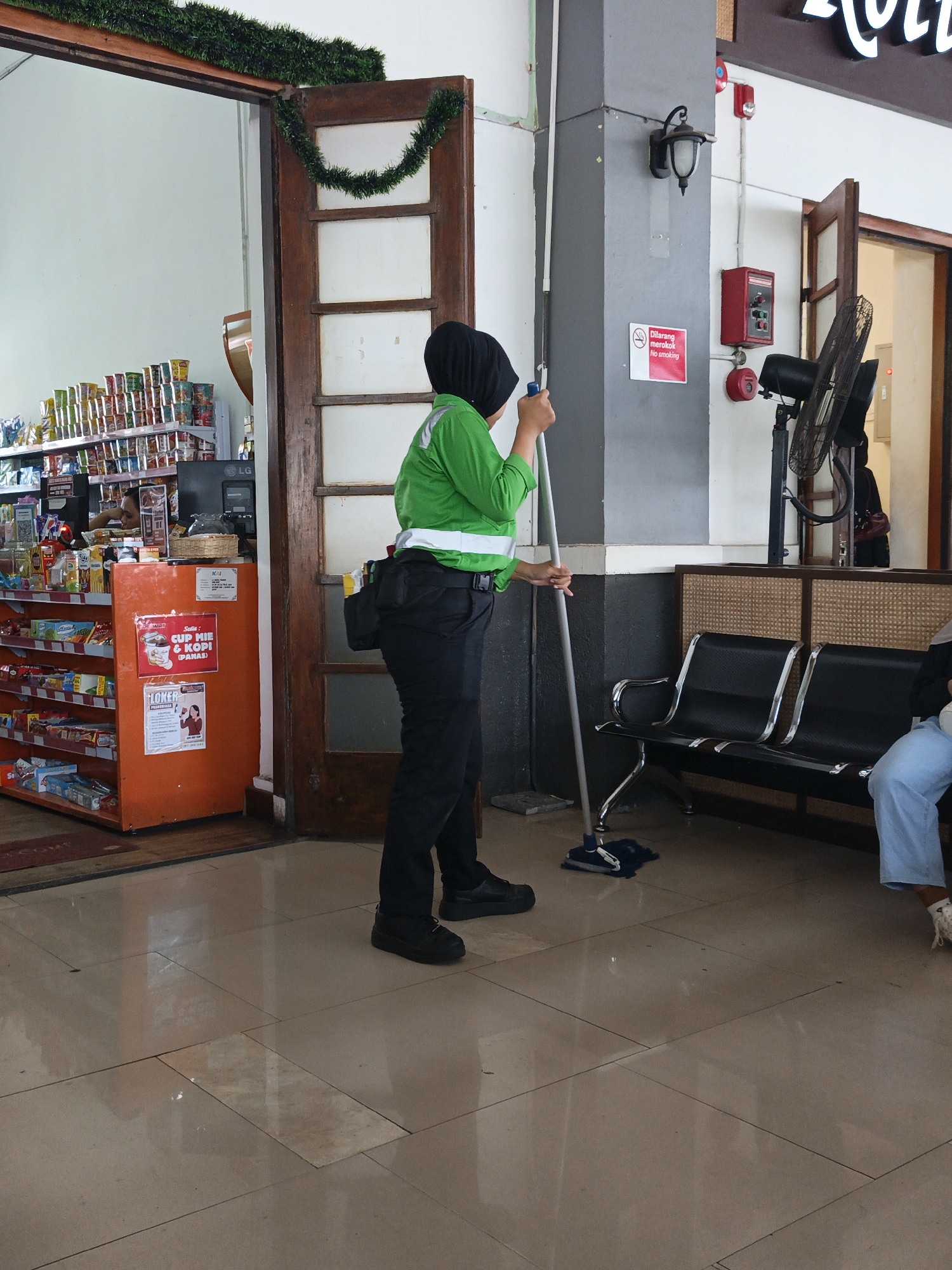 In Klaten, Indonesia, a person wearing a green jacket and black pants, along with a black headscarf, is mopping a tiled floor. The person is holding a mop with a blue mop head and a silver handle. The environment appears to be an indoor area with shelving units stocked with various products, possibly a small store or convenience store, visible to the left of the image. A wooden doorframe is also present. In the background, there are black seats and a standing fan. A red fire alarm box and a "No Smoking" sign are affixed to a pillar. The overall scene suggests a public indoor space undergoing cleaning.
