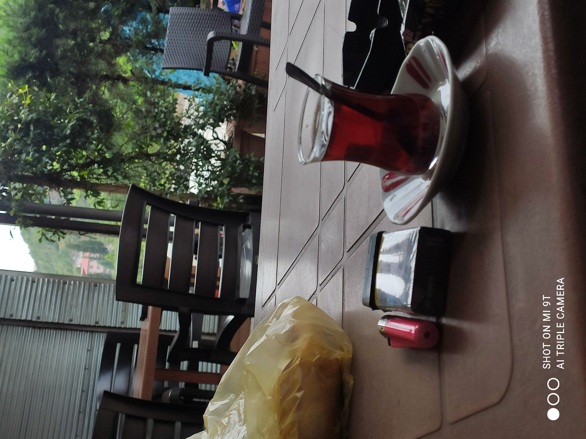 A close-up, eye-level, outdoor shot captures a table setting at what appears to be a cafe in Akçaabat, Turkey. The table, made of a brown, patterned material, holds several items. In the foreground, a red lighter and a pack of cigarettes are placed near a glass of dark red liquid, presumably tea, with a spoon. The glass sits on a saucer with a white and red patterned rim. To the left, a crumpled yellow plastic bag is visible, suggesting food or goods. In the background, dark wooden chairs and a table are seen, hinting at a seating area. Beyond that, a corrugated metal wall partially obscures a view of lush green trees and foliage, with glimpses of buildings nestled within the landscape, indicative of a semi-urban or suburban setting. The overall impression is a casual outdoor break.