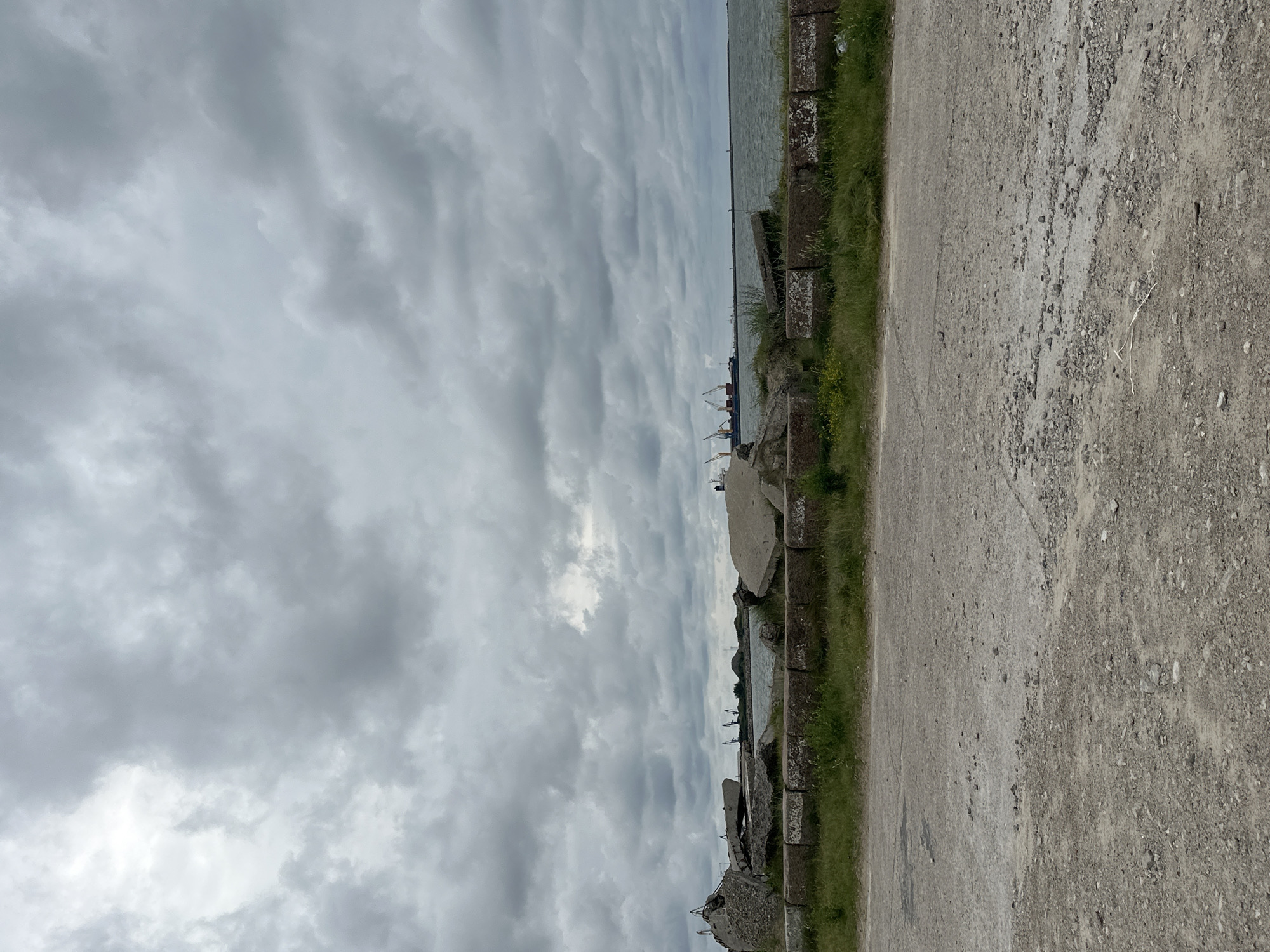 The image depicts a cloudy day at what appears to be a port or industrial area, likely in Liepaja, Latvia, given the context provided. In the foreground, a rough, gravelly path runs horizontally across the frame. Behind the path, a strip of green grass is bordered by a line of large, weathered concrete blocks, some of which show white markings. Beyond the blocks and grass, the scene opens up to a body of water, possibly a bay or sea, with choppy waves.
On the far side of the water, a harbor is visible with several large cargo ships docked. Towering cranes, typical of port operations, are prominent. The sky is overcast with heavy, gray clouds, creating a somber atmosphere. Scattered concrete debris is present along the shoreline in the mid-ground, suggesting a site that might be under construction, demolition, or has experienced damage. No people are visible in the image. The overall impression is one of a working port under a dull sky.
