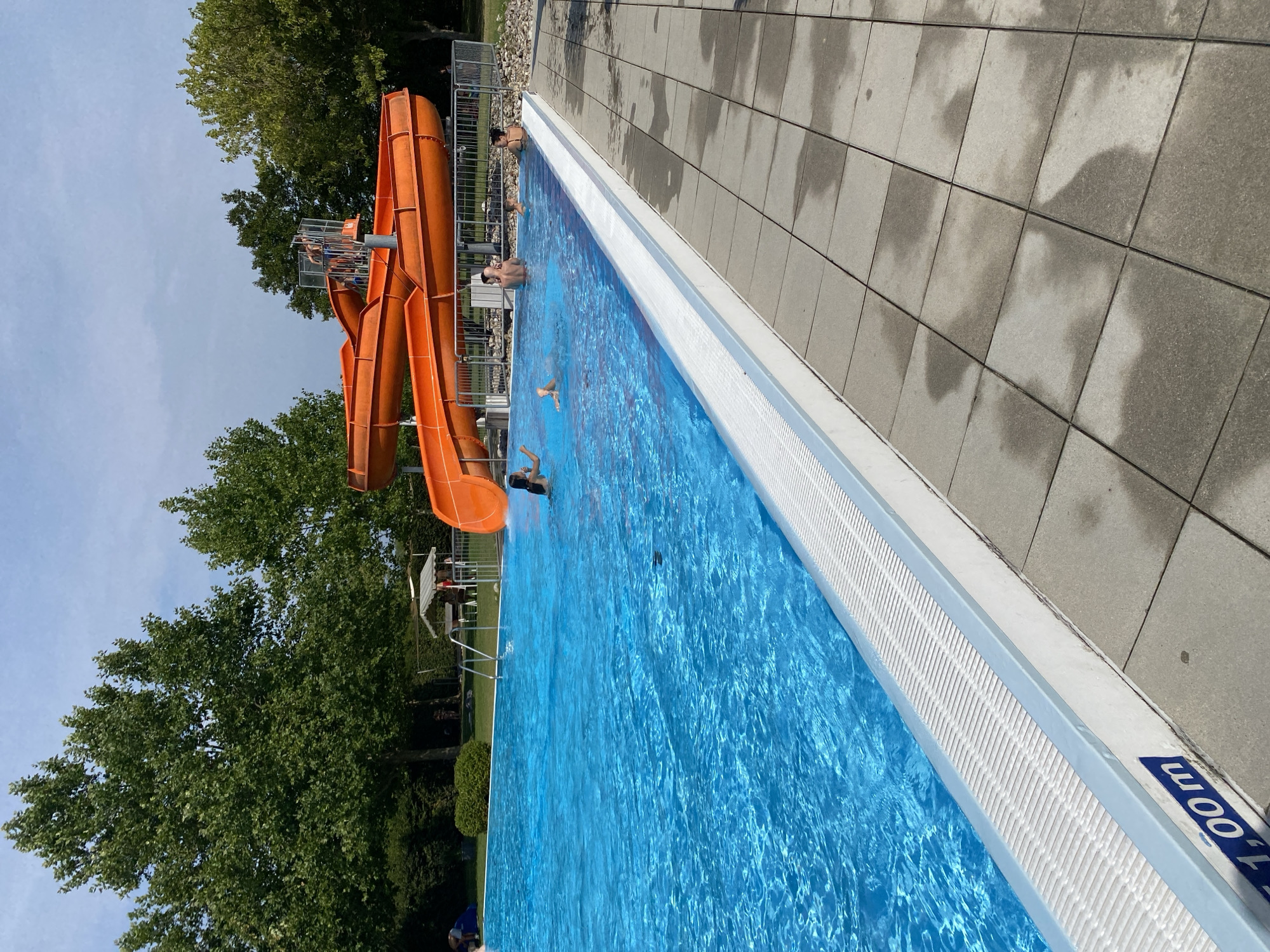 The image captures a bright, sunny day at an outdoor swimming pool complex, likely the public pool in Deutschkreutz, Austria, given the presence of a "1,00m" depth marker. The pool itself is a vibrant, clear blue, with several people visible enjoying the water. In the foreground, the edge of the pool features a white, textured overflow channel and a concrete patio tiled with grey squares, some of which are wet.
Dominating the background and to the right are two large, interconnected orange water slides. A metal railing surrounds the base of the slides and the top platform where a few individuals can be seen. Lush green trees form a dense backdrop to the entire scene, with a clear blue sky overhead. A few picnic tables with a canopy are visible on the grassy area behind the pool. The overall impression is of a lively and recreational public space.