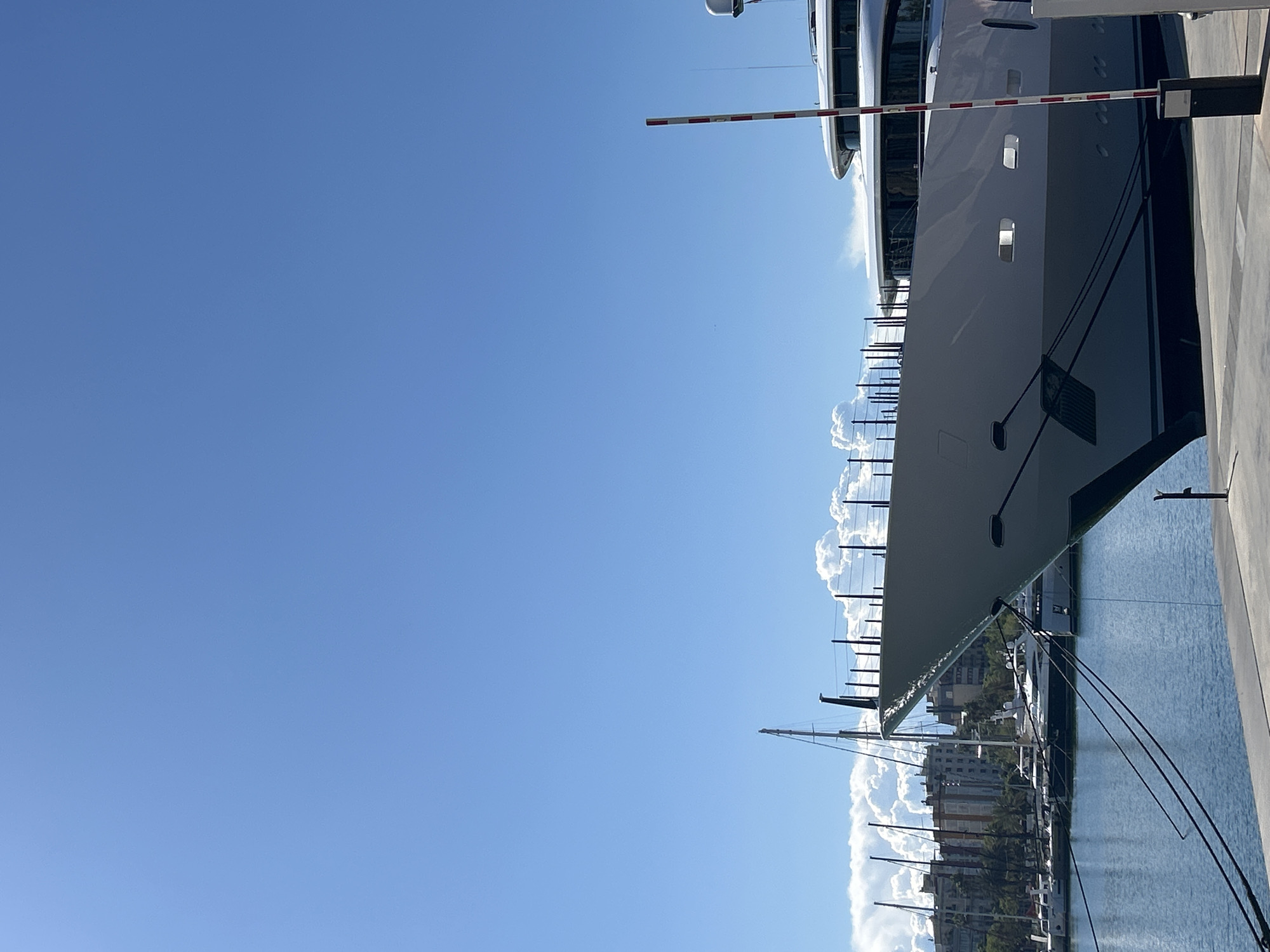 The image shows a view from ground level in a marina in Barcelona, Spain. A large, modern yacht dominates the right side of the frame, docked alongside a concrete pier. The yacht is gray with dark accents and has multiple windows. A red and white striped barrier arm is positioned above part of the yacht. A few other sailboats and yachts are visible in the background of the marina. Some city buildings are also visible in the background. The water is calm, reflecting the sky. The sky is clear and blue with some wispy clouds near the horizon.