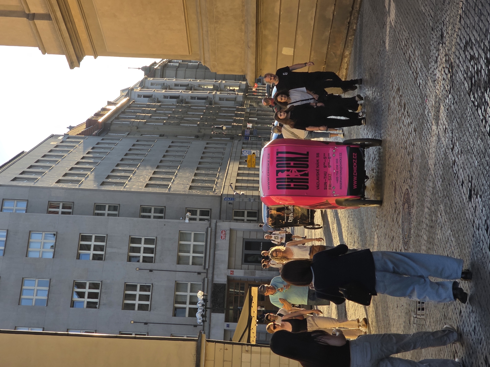 The image captures a bustling street scene in a European city, likely Prague, under the warm glow of late afternoon or early evening sunlight. The cobblestone street is lined with multi-story buildings featuring numerous windows and classic architectural elements.
In the foreground, several individuals are walking or standing, their faces partially obscured or turned away. One person prominently featured wears a black jacket and light blue jeans. Further into the scene, a group of people congregates around a vibrant, bright pink three-wheeled cart or mobile vendor. Two men in dark clothing are visible near this cart.
The pink vehicle is a key focal point, adorned with text advertising "CHICKZ.CZ". Visible details on the cart include "VÁCLAVSKÉ NÁM. 56" (Wenceslas Square 56), operating hours "SUN-THU 9PM-5AM" and "FRI-SAT 9PM-6AM", a phone number "+420 732 232 333", and an email address "RESERVATIONS@CHICKZ.CZ". The website "WWW.CHICKZ.CZ" is repeated. On a building in the midground, a sign partially reads "FRANZE KAFKA," strongly suggesting the location is Prague.
The activity appears to be casual street life, with people moving about or interacting. The long shadows and warm light indicate a clear, sunny day. The overall atmosphere is typical of an urban environment during leisure hours.
