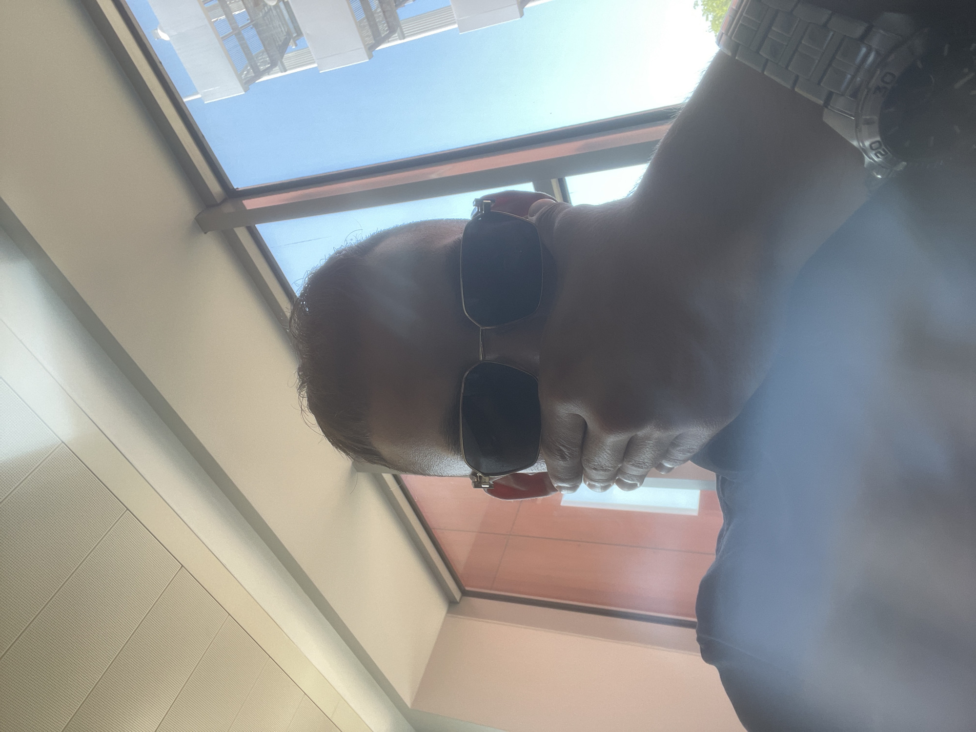 A person's face is obscured by their hand and sunglasses as they are photographed from the chest up, indoors, near a window with a view of a blue sky and a modern building. The person is wearing a dark shirt and a silver watch on their left wrist. The background includes a light-colored ceiling with panels, a wall with a reddish-orange hue, and a window frame. The lighting suggests it is daytime, with sunlight coming through the window.