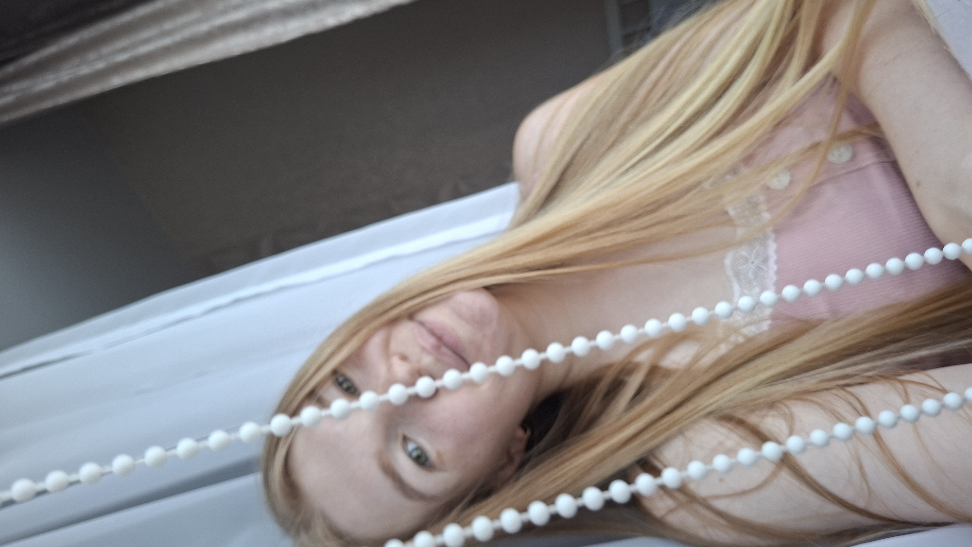 This is a close-up, rotated portrait of a young woman with long, blonde hair. She is wearing a pink top with white lace trim and white buttons. A string of white beads runs across the foreground, obscuring parts of her face and upper body. Her eyes are green and she is looking towards the camera with a soft, slightly amused expression. The background appears to be an indoor setting, possibly a room with light-colored curtains or blinds. The lighting suggests it could be daytime, but the exact time of day or weather conditions are not discernible. There is no visible text in the image. The setting appears to be domestic.