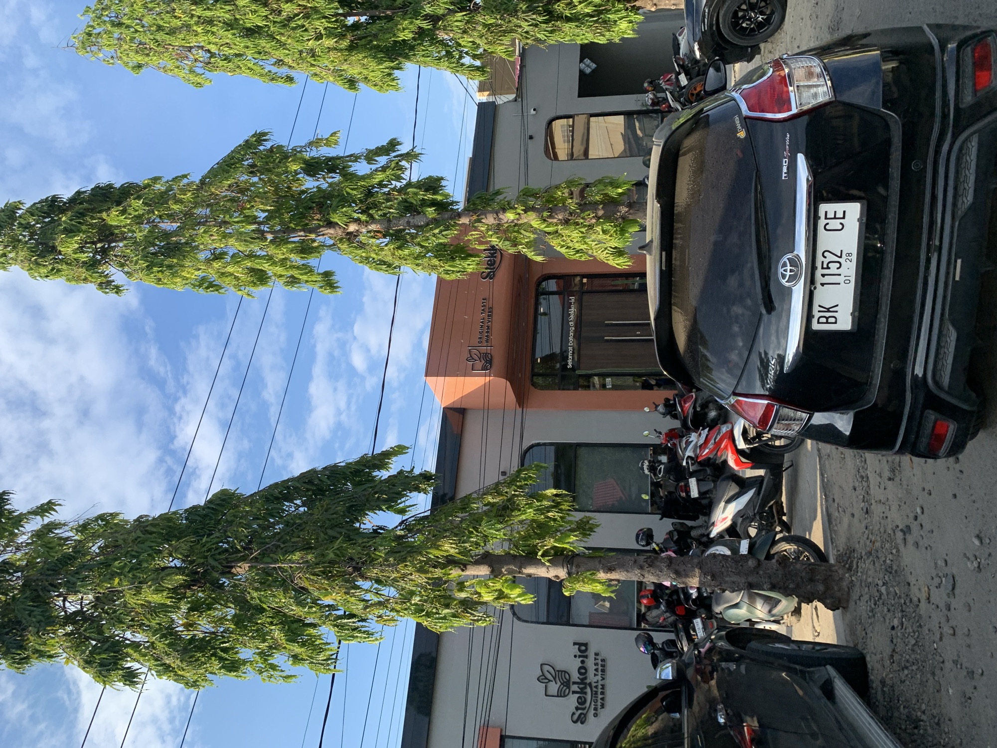 The image depicts the exterior of a building identified by the logo "Stekko.id" and the tagline "ORIGINAL TASTE WARM VIBES". The building has a modern design with large windows and an orange accent section. In front of the building, a parking area is visible, filled with several motorcycles and two cars, one of which is black and prominently features a license plate that reads "BK 1152 CE". The scene is set in a city environment, likely Lhokseumawe, Indonesia, indicated by the license plate format. Tall, slender trees with lush green foliage line the area, and the sky above is blue with scattered white clouds. The ground is covered with gravel. There are no people directly visible in the image, suggesting the scene was captured at a time when the establishment might be closed or patrons are inside.
