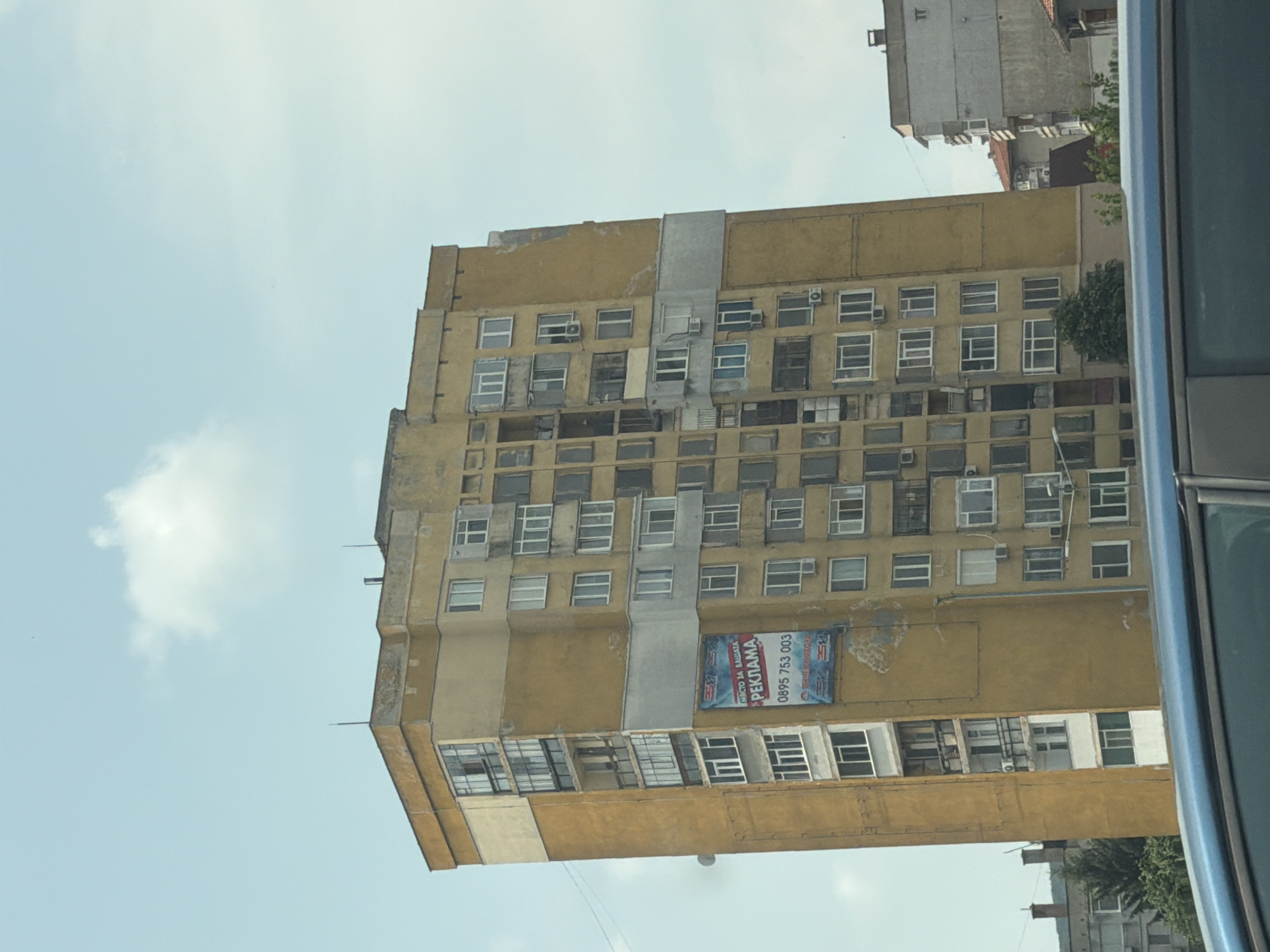 This image captures a tall, aged, yellow-ochre building against a bright, light blue sky with scattered white clouds. The photograph appears to be taken from a slightly upward angle, with the right edge of the frame showing a blurred portion of a blue vehicle, suggesting it was captured from inside a car.
The main subject is a multi-story building, likely residential or commercial, characterized by its faded yellow facade and numerous rectangular windows. Many windows are either covered, appear vacant, or have external air conditioning units attached. The architectural style suggests an older construction. Patches of lighter grey cement are visible on the building's exterior, indicating repairs or additions.
A large blue and white banner is prominently displayed on the side of the building. The text on the banner is in Cyrillic script and reads "МЯСТО ЗА ВАШАТА РЕКЛАМА", which translates to "PLACE FOR YOUR ADVERTISEMENT". Below this, a phone number "0895 753 003" is clearly visible, indicating the availability of advertising space.
In the background, beyond the yellow building, another lighter-colored building and some green foliage are partially visible. The scene is static, with no discernible activity or people present. The bright sky indicates it is daytime. The image orientation is rotated about 90 degrees clockwise from a conventional upright view. The context places this scene in Shumen, Bulgaria.