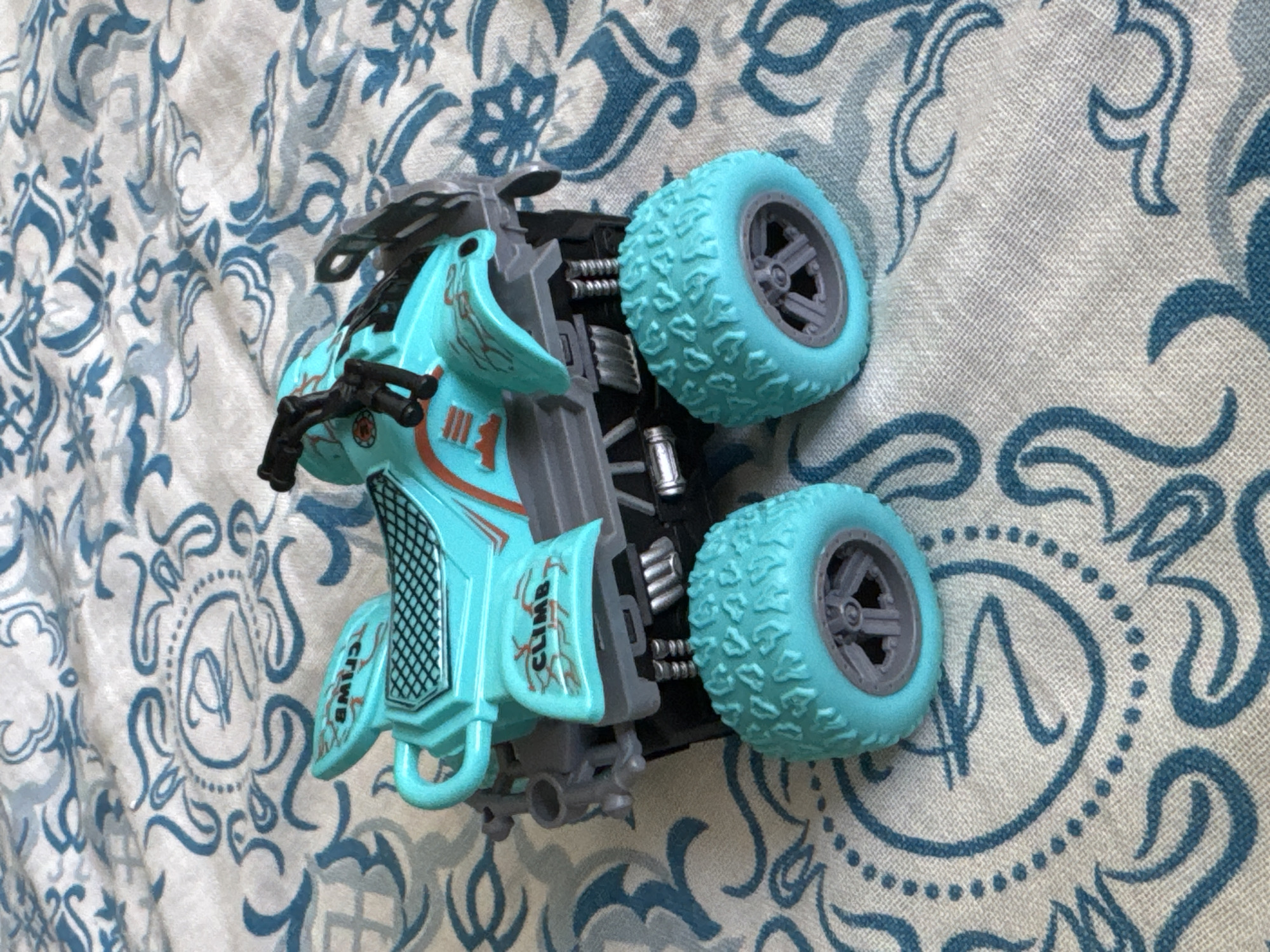 A turquoise toy ATV with grey accents and black handlebars is displayed on a white and blue patterned fabric. The ATV has the word "CLIMB" printed in white on one of its side panels. The fabric has an ornate pattern with swirls and circular designs. The image does not contain any information about the location in the city of Яромінскі сельскі Савет, Belarus.