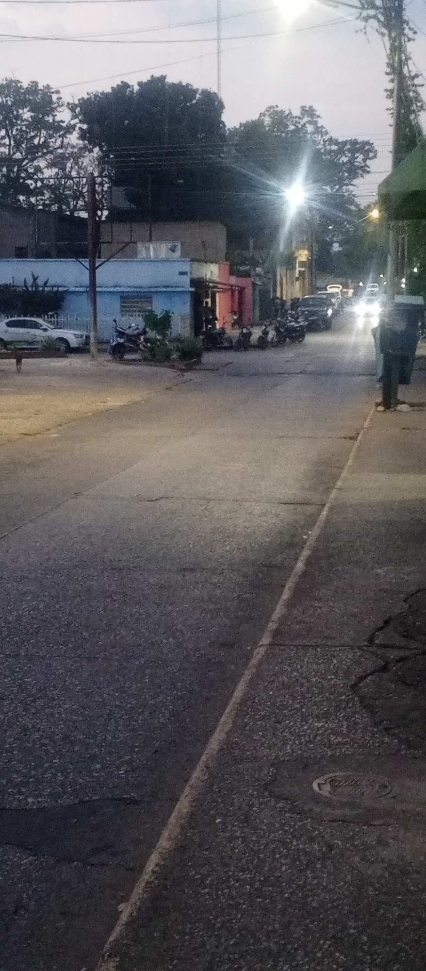 This image shows a street scene in Tinaquillo, Venezuela, at dusk or twilight. The street is asphalt, with visible cracks and a curb along the right side. A row of parked motorcycles lines the street near a building with light blue and pink facade elements. Several cars with illuminated headlights are visible further down the street. Trees are present in the background and along the sides of the street. Streetlights are on, casting a bright glow. A trash bin stands on the sidewalk to the right. A white car is parked on the left side of the street.