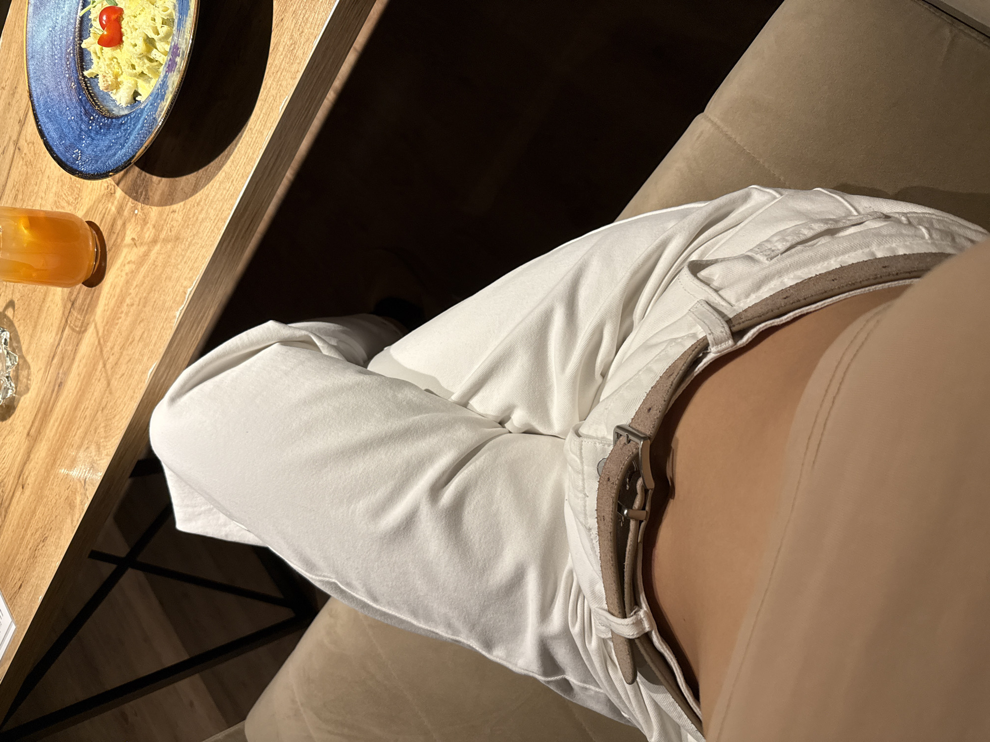 The image shows a first-person, close-up view of a person seated at a wooden table. The person is wearing white trousers secured with a brown leather belt. The waist and part of the abdomen are visible. On the table, there is a blue plate containing pasta with a cherry tomato on top, and a glass of orange juice. The background shows a dark area and a light-colored couch or seating surface. There is no explicit information provided to indicate the location is in Chaikovsky, Russia.