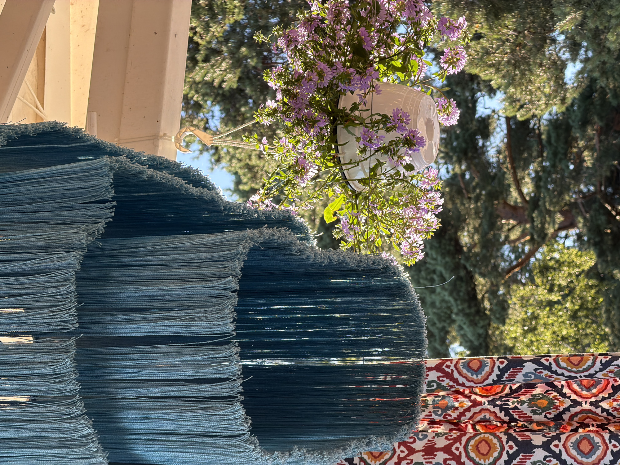 The image depicts an outdoor scene with a focus on decorative elements and nature. In the foreground, a large, multi-tiered chandelier made of teal-colored fringe hangs from a white overhang, likely part of a porch or gazebo. To the right, a white hanging planter filled with small, pale purple flowers is suspended by a rope and hook, also attached to the overhang. The planter is positioned beneath a cluster of the chandelier's fringe. A section of patterned fabric with a bohemian-style geometric design in red, orange, blue, and white is visible on the left side of the frame, partially obscured by the fringe. In the background, lush green pine trees are visible under a clear blue sky, suggesting a natural, possibly wooded, environment. The lighting indicates it is daytime, with sunlight casting highlights and shadows. The scene is set outdoors, possibly in a garden or on a patio, with elements of decor and vegetation. This specific location in the city of Komarovo, Russia, is a dacha or residential area characterized by its proximity to nature and its use of outdoor decorative elements.
