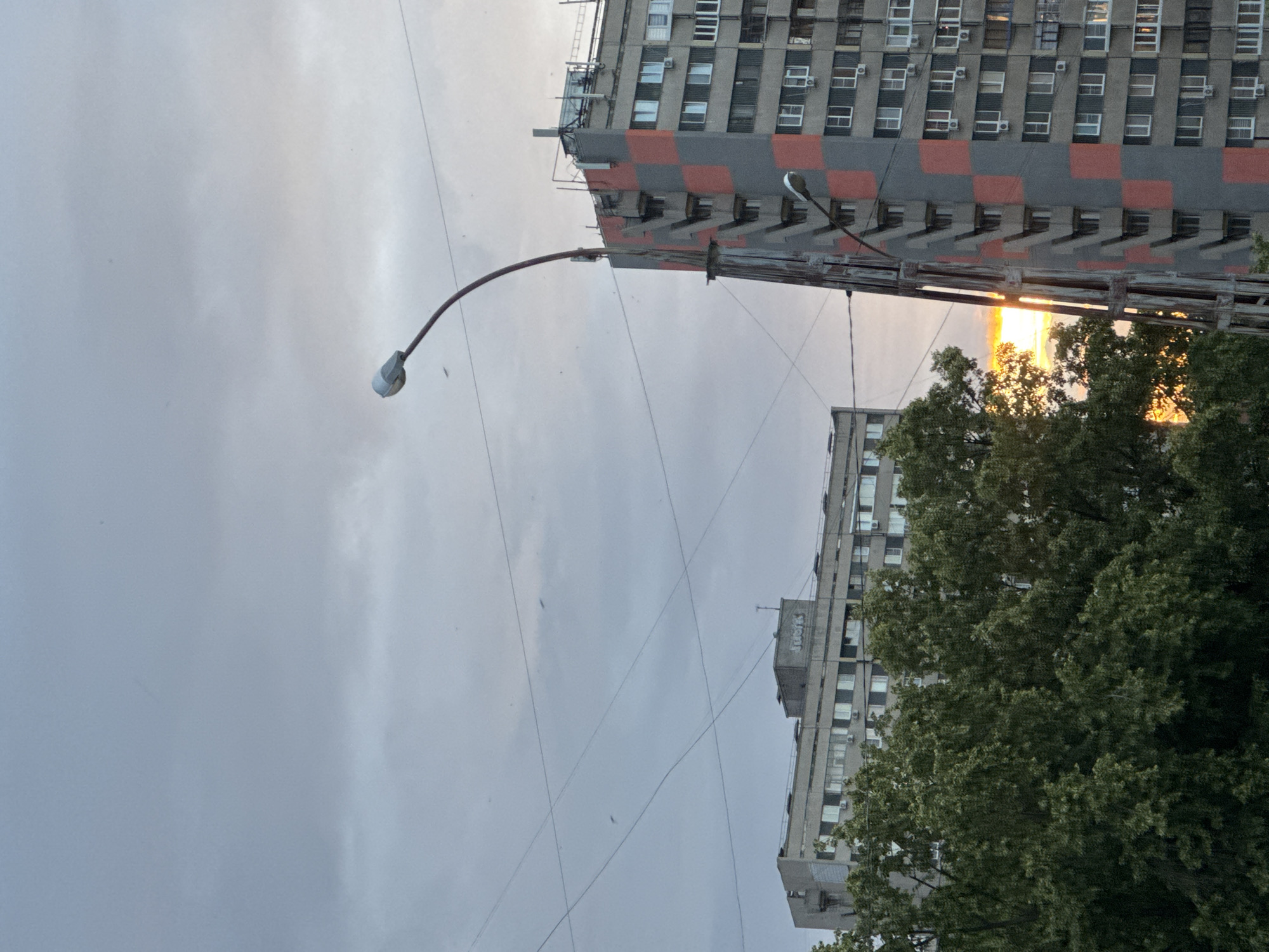 An exterior, medium shot captures a sky with scattered clouds and a sliver of sunset peeking through. In the foreground, a tall, curving lamppost extends towards the sky on the right. To the left of the lamppost and further back, a row of trees with dark green foliage is visible. Behind the trees and lamppost, two multi-story buildings dominate the scene. The building on the right is taller and features a grey facade with a checkerboard pattern of red and grey squares. The building on the left is lower and has a plain grey facade. Power lines crisscross the sky between the buildings. The overall lighting suggests late afternoon or early evening.