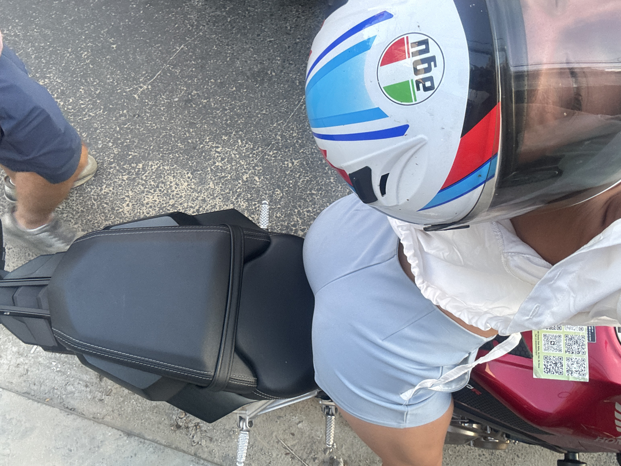 A high-angle shot shows a person wearing a white AGV motorcycle helmet, light blue shorts, and a white top, sitting on a red Honda motorcycle. The motorcycle has a black seat. A person wearing dark shorts and silver shoes stands to the left of the motorcycle. The scene is set in a street or parking area within Hội An Ward, Vietnam.