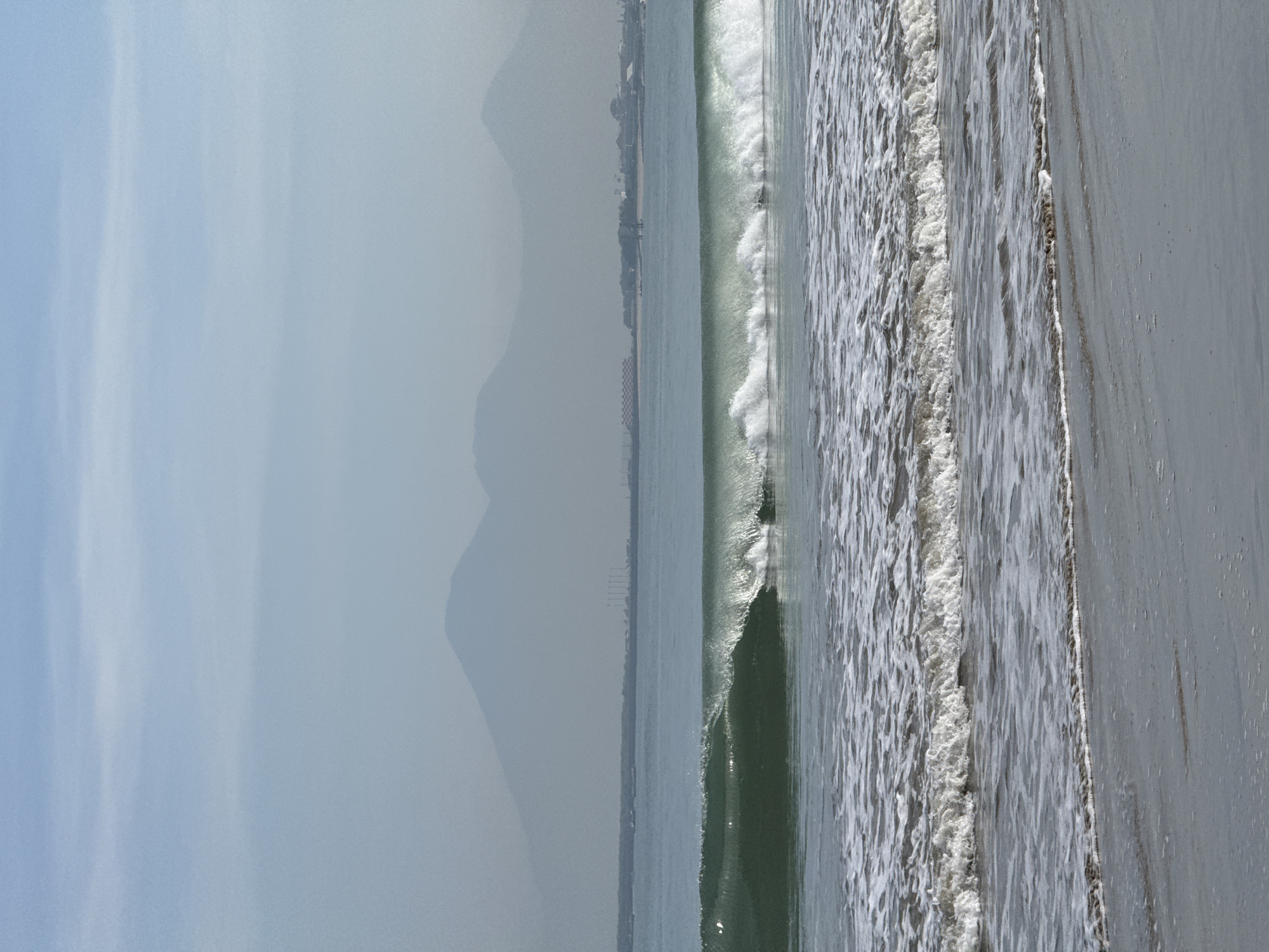 The image shows a beach scene, likely in Kuta Selatan, Indonesia. The photograph is oriented vertically. The foreground consists of a sandy beach with visible wet sand closer to the water. Waves are crashing onto the shore, with foamy white water visible. In the distance, a hazy coastline is visible, with mountains obscured by atmospheric conditions in the far background. There are signs of human habitation along the distant coast. The sky is overcast, contributing to the hazy atmosphere. The focus is on the beach and the wave action. There are no people visible in the image.