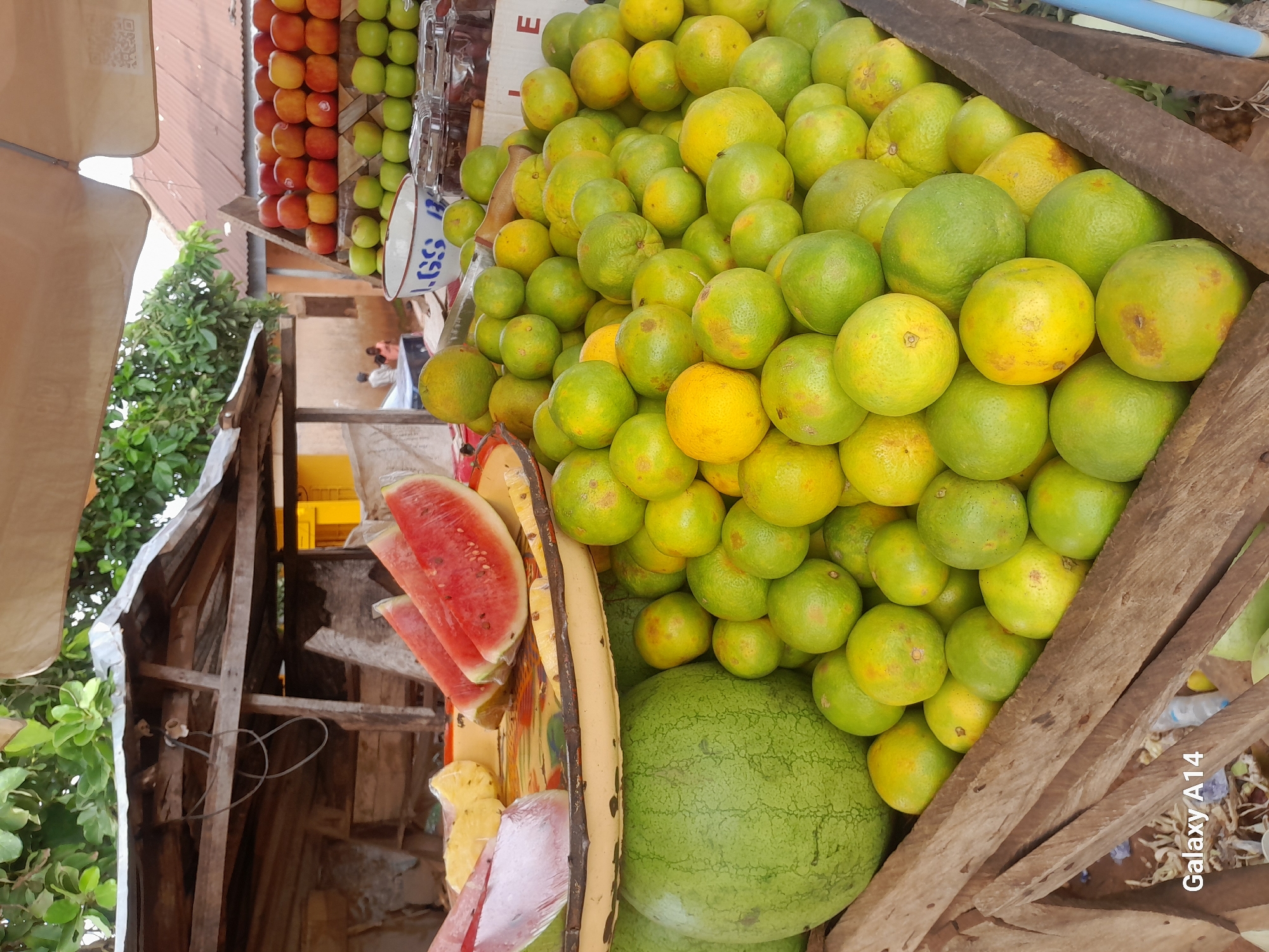 The image depicts a fruit stall in Barakin, Nigeria. A variety of fruits are displayed, including a large watermelon, sliced watermelons on a platter, pineapples, and numerous oranges or tangerines piled in wooden crates. Behind the fruit stall, a rustic wooden structure with a corrugated metal roof is visible, suggesting an outdoor market setting. In the background, a few individuals can be seen, and the overall environment appears to be an outdoor marketplace with simple structures. The photograph is taken from a slightly elevated angle, looking down at the fruit display.