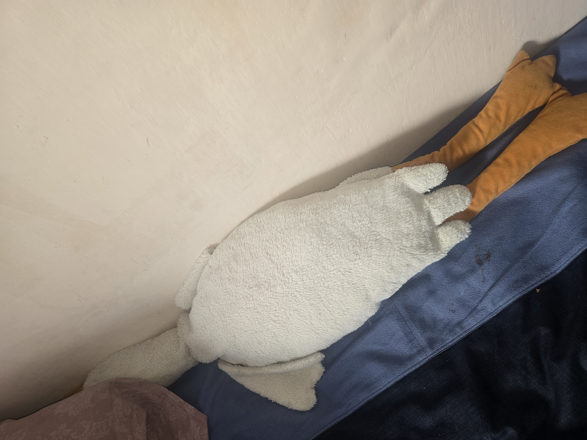 The image shows a stuffed toy goose lying on a blue surface, possibly a bed or couch. The goose is white and fluffy, with orange legs and feet. A pinkish-brown patterned fabric is visible behind the goose's head, and a pale pink wall is behind that. The scene is indoors, likely in a residential setting. There is no discernible information to indicate its location in Ulan-Ude, Russia.