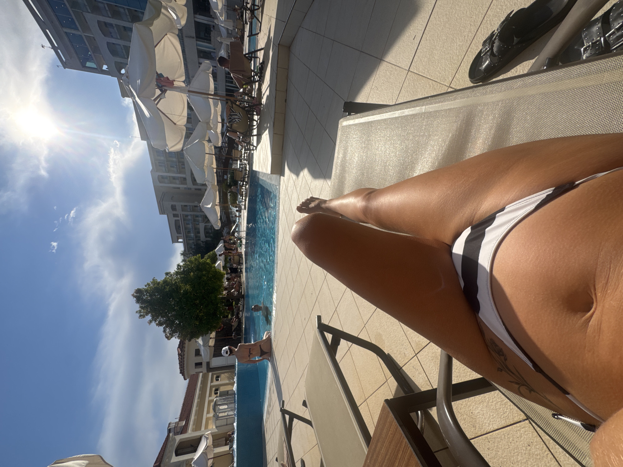 The image depicts a sunny day at a resort pool area, likely in Boreti, Montenegro, given the context. In the foreground, a person is lying on a lounge chair, with their legs and torso visible, wearing a black and white striped bikini. Their legs are tanned and positioned slightly upward. To the right of the chair, a pair of black slides rests on the tiled ground.
The midground features a large swimming pool with blue water. Several people are in and around the pool. Two individuals are partially submerged in the water near the edge. Further back, people are seated on lounge chairs under white umbrellas, some appear to be reading or relaxing. A green tree stands prominently near the pool.
The background showcases a multi-story hotel building with modern architecture. The sky is a bright blue with scattered white clouds, and the sun is shining intensely, creating lens flare in the upper right portion of the image. The overall environment suggests a recreational setting, possibly a hotel or resort, with a focus on leisure activities by the pool.