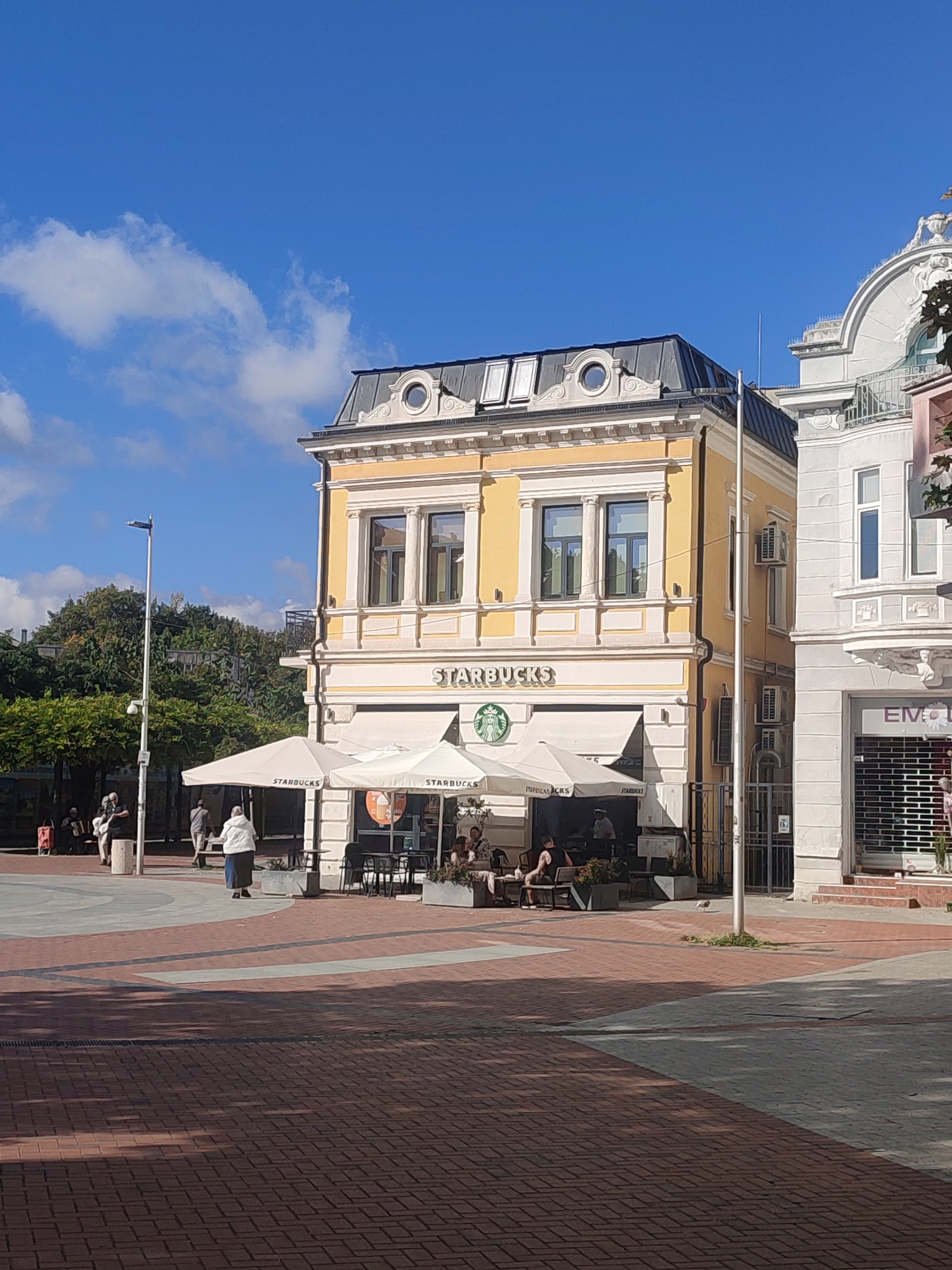 The image depicts a sunny day in the city center of Varna, Bulgaria, near the Cathedral of Saint Mary. In the foreground, a red brick pedestrian area is visible with dappled sunlight and shadows cast by trees. A light gray path intersects the brickwork. Several people are scattered throughout the scene. In the mid-ground, a Starbucks cafe is prominently featured. It has a yellow facade with white architectural details, large windows, and an outdoor seating area with white umbrellas bearing the Starbucks logo. People are seated at tables under the umbrellas, and one person is standing near the entrance. To the right of the Starbucks, another building with a white facade and arched windows is partially visible, with a shop entrance covered by a metal grate. Further to the left, a street lamp and lush green trees form the backdrop. In the far background, a clear blue sky with a few white clouds completes the scene.