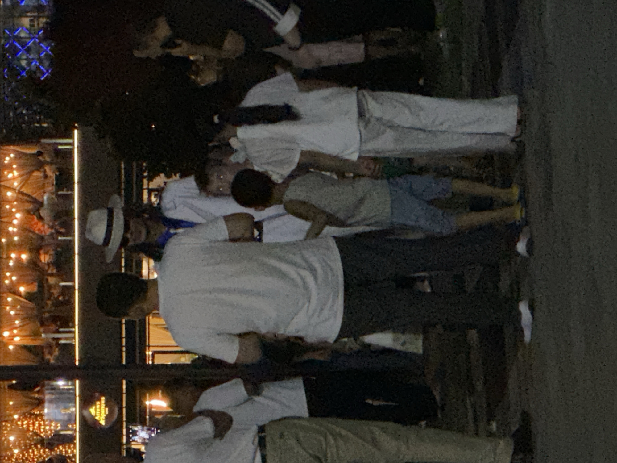 The image depicts a group of people standing on a paved surface, likely outdoors at night. In the background, a building with illuminated windows is visible, with a sign that reads "Arrows & Sparrows." The lighting in the background suggests a vibrant or festive atmosphere, possibly a restaurant or entertainment venue. Several individuals are present, including adults and children. One person is wearing a white hat and a blue and white shirt, suggesting a distinct style. The attire of others varies, with some in casual clothing. The overall scene appears to be a social gathering or a moment captured in a public space. The presence of the "Arrows & Sparrows" sign and the visual context of urban nightlife suggest this scene could be located in the city of Tashkent, Uzbekistan, where such establishments are common.