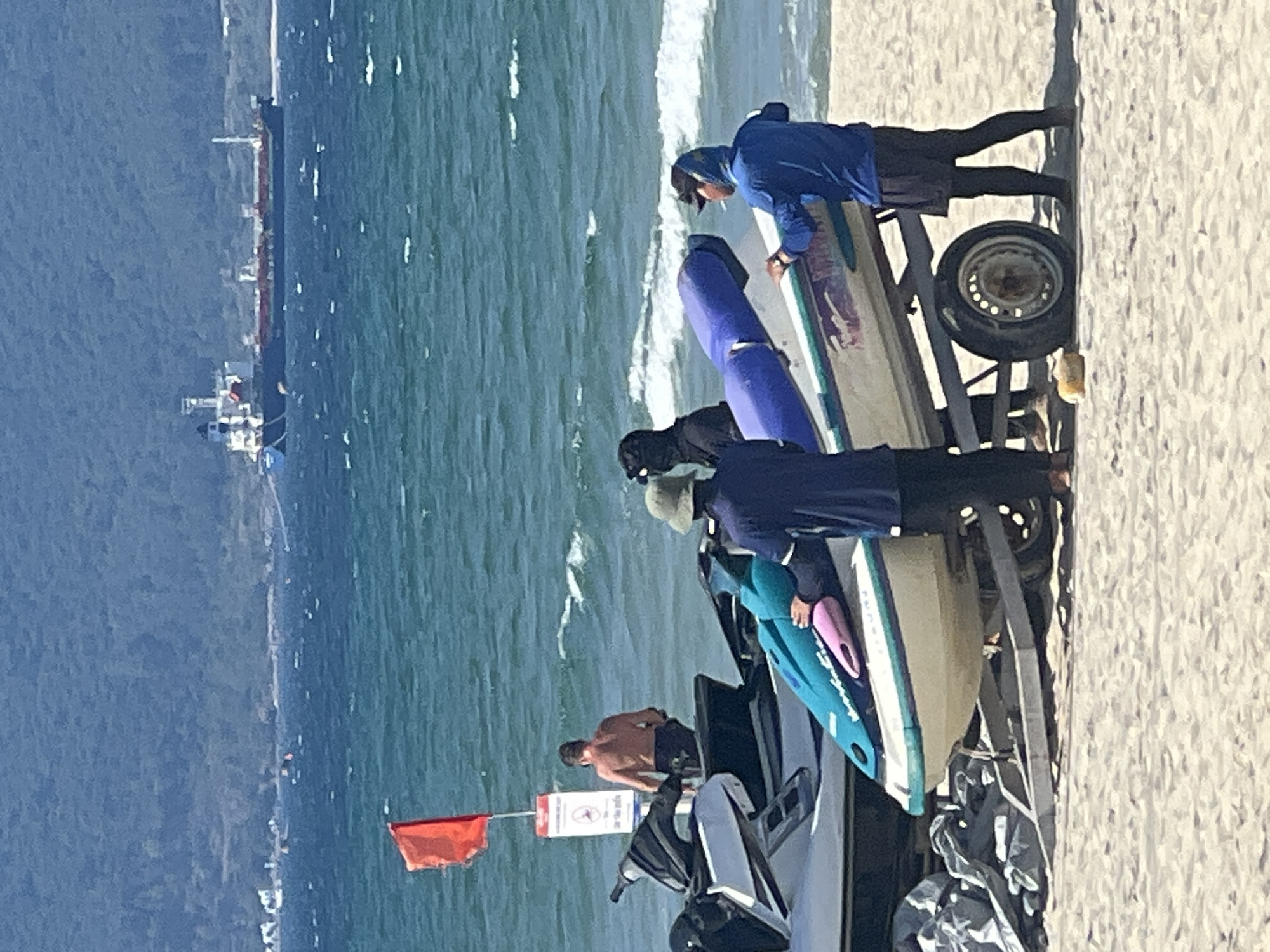 The image shows a sunny beach scene in Da Nang, Vietnam. In the foreground, a group of people are preparing to launch jet skis. Two jet skis are visible, one on a trailer and another partially covered with a tarp. Three individuals are interacting with the jet ski on the trailer; one person wearing a blue hoodie and black shorts is standing on the right, appearing to smoke a cigarette and push the jet ski forward. Another person in a blue shirt and khaki hat is to the left, holding a blue and pink flotation device. A third person, wearing a black face covering, is behind the jet ski. In the background, a shirtless man stands near the water's edge, and a red flag is planted on a pole. The ocean is a deep blue with moderate waves. In the far distance, a large cargo ship is visible on the horizon, with a lush green hillside behind it. The sand on the beach is light brown.