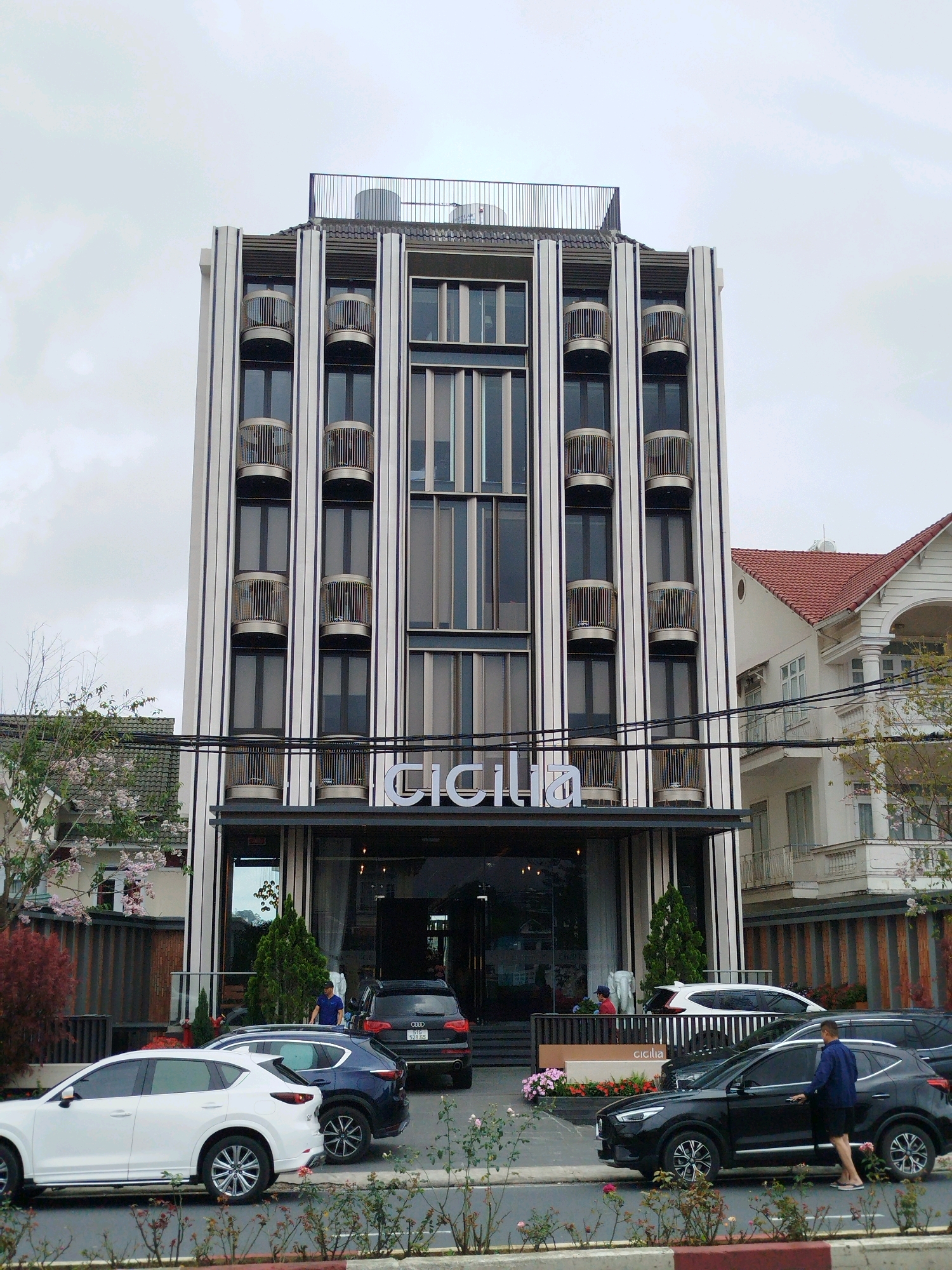 The image shows a modern, multi-story building with a distinctive facade featuring vertical white and dark grey stripes, punctuated by numerous small, circular balconies. The building has a prominent sign with the word "CICILIA" in large white letters above the entrance. In front of the building, a street with cars is visible. A white SUV and a dark blue SUV are parked on the left side, with a black sedan on the right. A man in a blue shirt and shorts is standing next to the black sedan, opening the driver's side door. A person in a red shirt is visible near the building's entrance. The sky is overcast. The building appears to be located in a developed urban area, possibly a city like Da Lat, Vietnam, given the architectural style and surrounding vegetation.