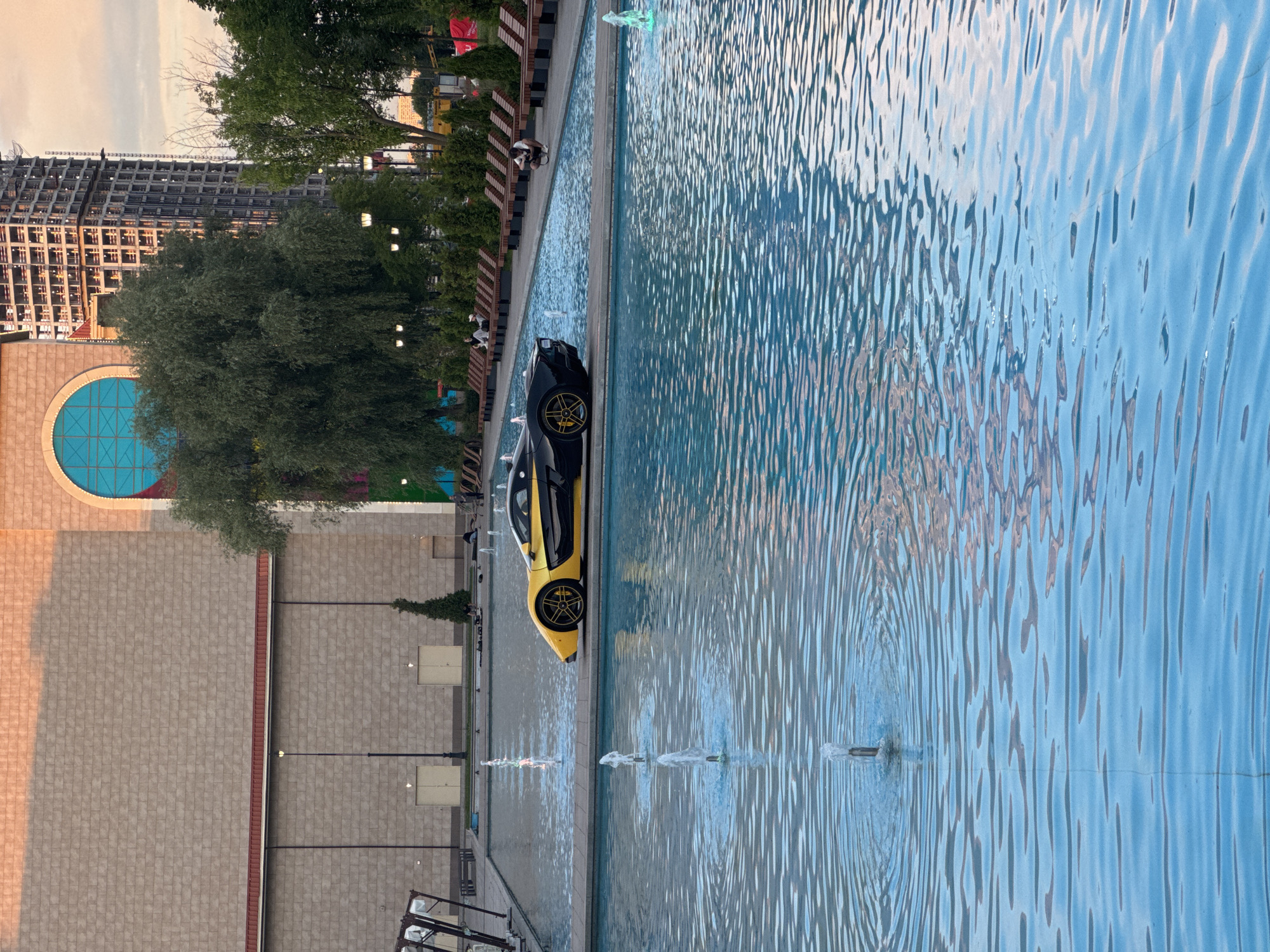A yellow and black sports car is parked on a raised concrete platform in the middle of a large, shallow fountain complex. The car is positioned horizontally across the platform, with its front facing left. The fountain has numerous spouts of water actively spraying. In the background, a large, tan, brick building with a red trim and a large arched window with a blue pane dominates the left side of the frame. To the right of the building, a tall, unfinished skyscraper under construction is visible. Green trees and park benches are arranged along the edge of the fountain complex. Several people are scattered throughout the scene, some sitting on benches and others standing. The sky above appears overcast with a hint of sunset colors. This scene is located at the "Muzeon" Art Park, adjacent to the Gorky Park, in Moscow, Russia.