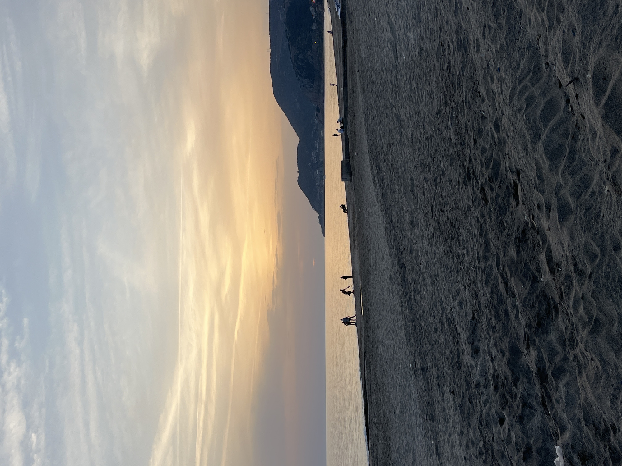 The image depicts a beach in Alanya, Turkey, at sunset. The sky transitions from light blue with wispy clouds on the left to orange and gold near the horizon. A mountain range is silhouetted against the sunset colors. The sea reflects the sunset hues. The beach is dark and sandy. Silhouetted figures of people are scattered along the shoreline.