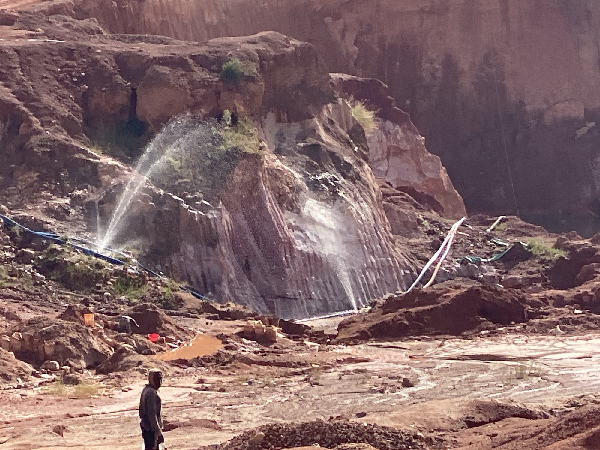 The image depicts an open-pit mining operation in a location identified as "Unknown, Unknown." The scene is characterized by reddish-brown earth and rock formations. Two water sprinklers are actively spraying water onto the rock face. A single person is visible in the foreground, standing near a pile of earth. In the background, there are buckets and what appears to be mining equipment. The environment appears muddy and disturbed.