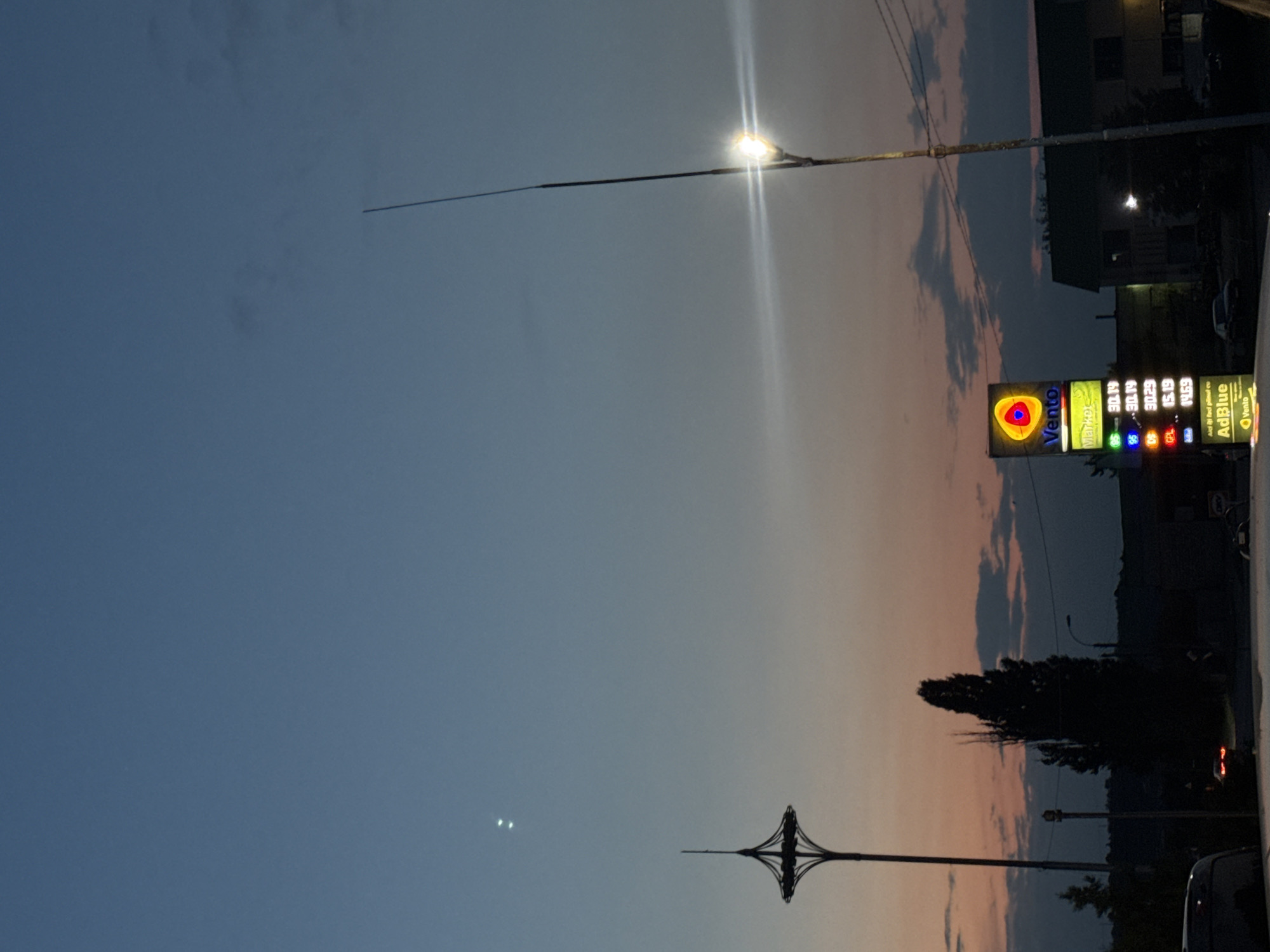 The image captures a dusky sky over Cimișlia, Moldova, with a vibrant gas station sign illuminated in the foreground. The sky displays a gradient of blues and oranges, with scattered clouds tinged pink by the setting sun. Two bright lights, possibly stars or distant lights, are visible in the upper left quadrant of the sky. A tall, slender structure with a pointed top stands to the left, and a light post with a bright bulb and lens flare is positioned to the right. The Vento gas station sign, featuring a colorful logo and prices for different fuel types (95, 98, Diesel, GPL, and AdBlue), is the central focus. Prices visible are 30.19, 30.29, 15.19, and 14.69. The sign is illuminated and stands vertically. Silhouettes of trees and buildings are visible in the background, along with a few vehicles, one with illuminated taillights. The overall scene suggests an evening or twilight setting in a semi-urban or suburban area.