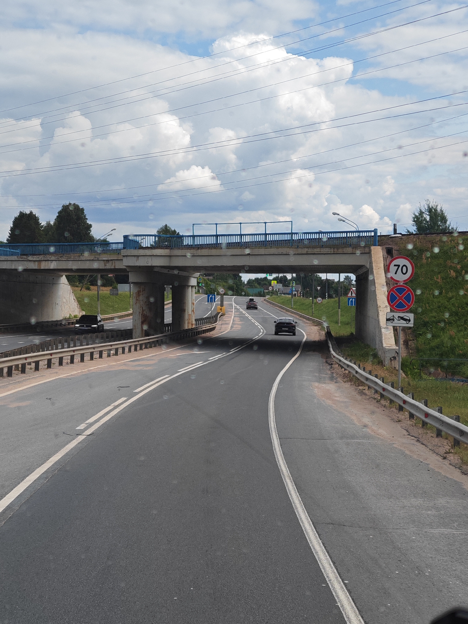 A view from a vehicle traveling on a two-lane asphalt road in Едрово, Russia. The road curves to the right and passes under a concrete overpass with a blue railing. On the right side of the road, there are traffic signs indicating a speed limit of 70, a no parking zone, and a tow-away zone. The environment is rural with trees and grass visible on the embankments. The sky is partly cloudy. Several cars are visible on the road, either approaching or moving away.