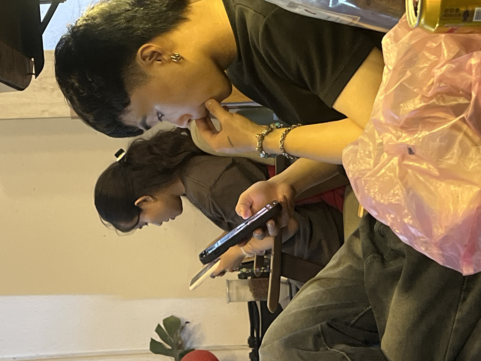 The image displays two young adults in an indoor setting, likely a cafe or restaurant, both engrossed in their smartphones. The photograph is oriented vertically, rotated 90 degrees clockwise from a natural upright perspective.
In the foreground, on the right side of the rotated image, a male individual is visible from the chest up. He has short, dark hair with closely shaved sides and a longer top. He wears a black t-shirt and multiple beaded and metallic bracelets on his left wrist. A distinctive multi-hoop earring is noticeable in his left ear. He holds a black smartphone in his right hand, while his left hand touches his chin or mouth, indicating deep concentration. To his right, a crinkled pink plastic bag and the top of a gold-colored beverage can with some unidentifiable text are visible.
Behind and to the left of the male, a female individual is partially seen. She has long, dark hair and wears a dark-colored top. She is also focused on a smartphone, holding it with both hands.
The background features a plain light-colored wall. In the lower left corner of the image, a plant with broad green leaves and a red spherical object (possibly a decorative pot) can be seen. A brown strap or handle is also visible near the female's phone.
The lighting is warm and somewhat dim, suggesting an evening or nighttime atmosphere. Both individuals appear relaxed and deeply absorbed in their digital devices. The scene is situated in Đà Nẵng, Vietnam.