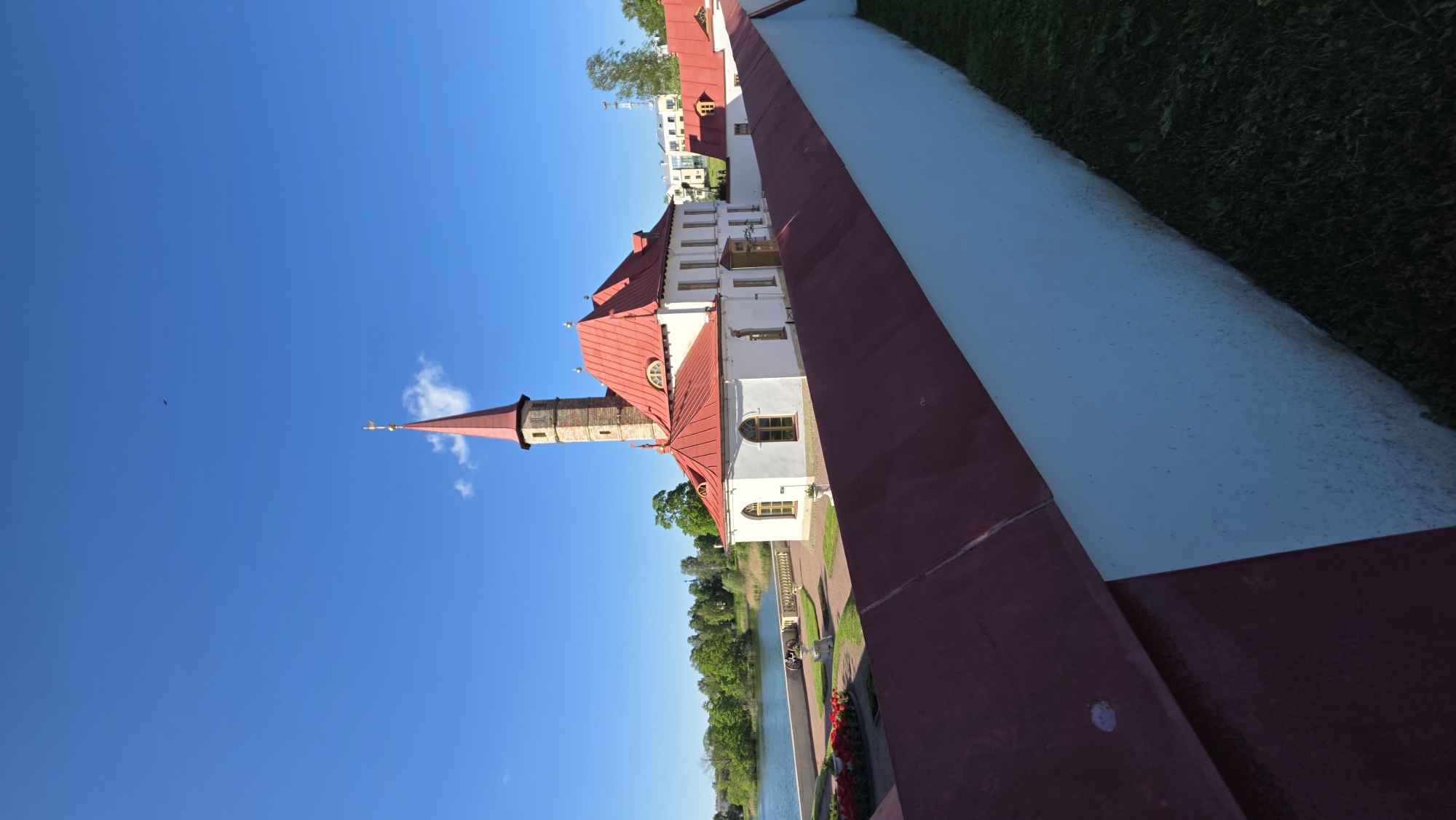 The image depicts the Prioratsky Palace in Gatchina, Russia. The palace is distinguished by its red roof and spire. The sky is clear blue with a few clouds. A body of water and greenery are visible in the background. A red and white painted structure in the foreground provides a sense of perspective.
